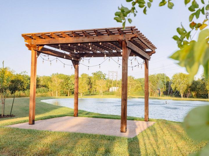 Wooden pergola with string lights on a concrete pad, overlooking a pond.