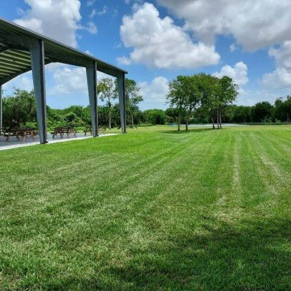 Lush green lawn with a covered metal structure and trees under a blue sky with clouds.