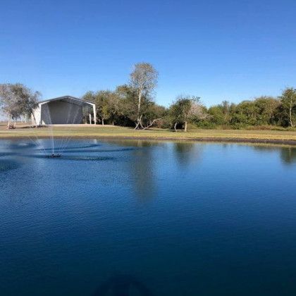 A blue pond with a fountain in front of a white building with trees in the background under a blue sky.