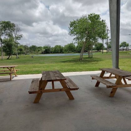 Picnic tables on a concrete patio overlooking a grassy field with trees and a small pond under a cloudy sky.