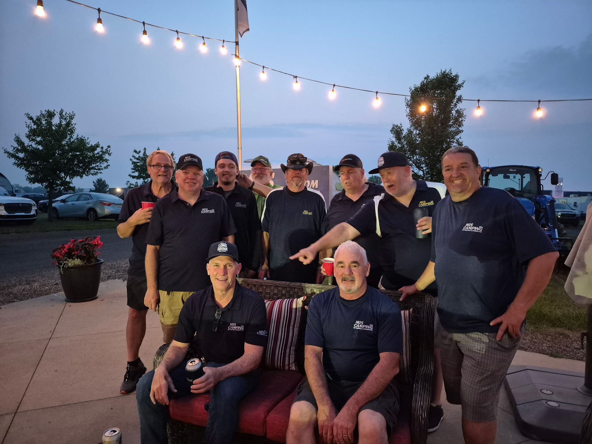 Group of men gathered outdoors at dusk, posing with drinks. Lit by string lights, near vehicles and flowers.