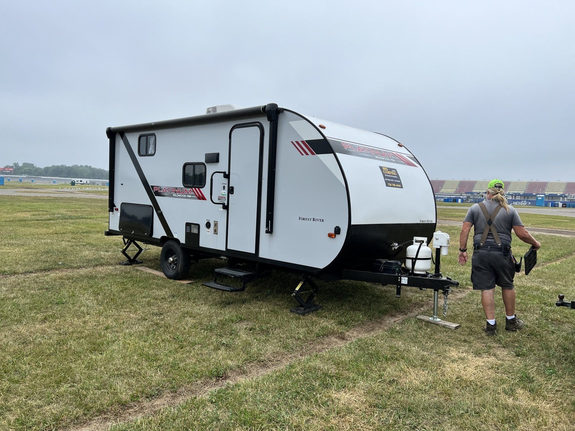A man is standing in front of a small trailer in a grassy field.