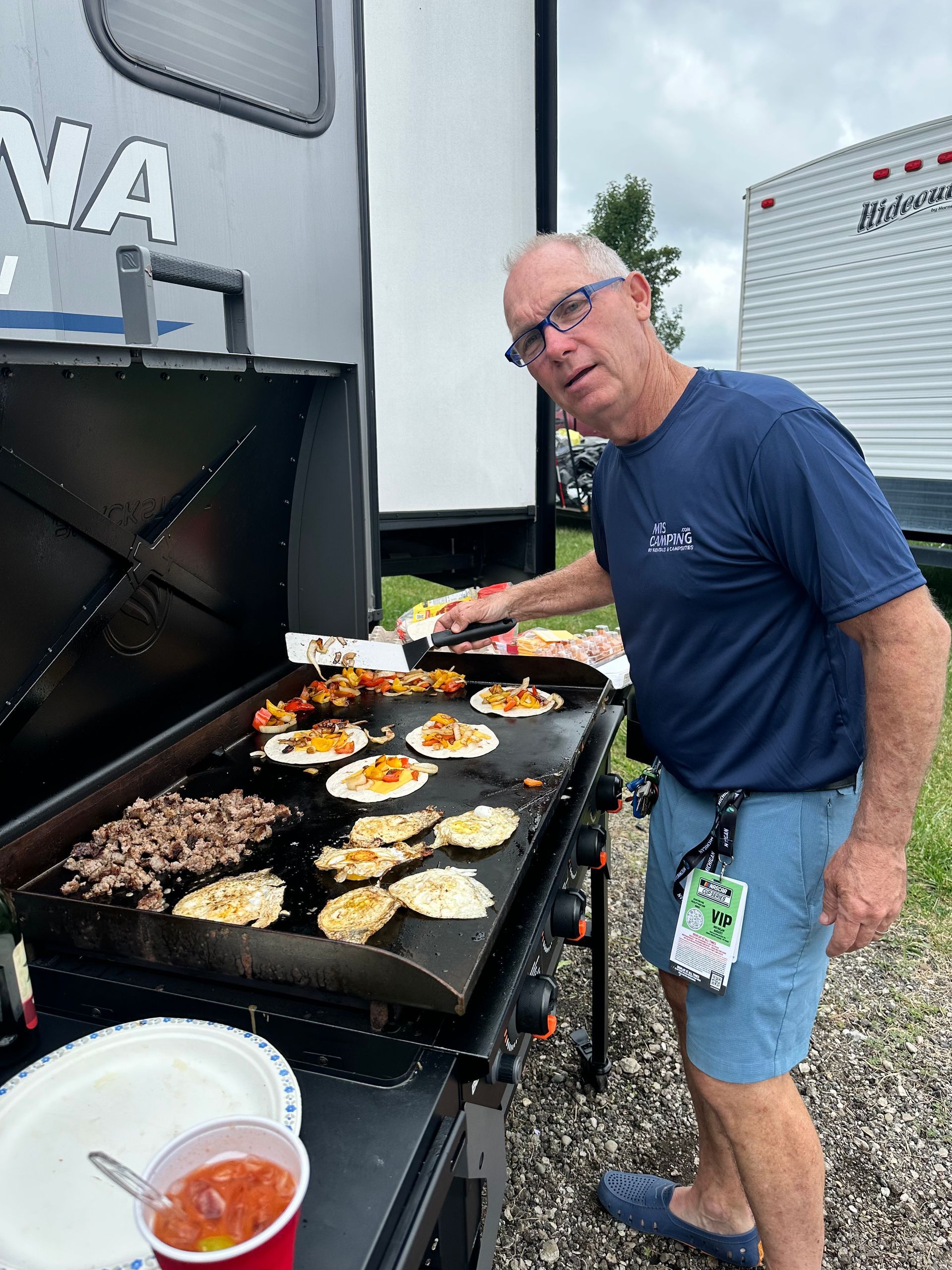 Man grilling food on a black griddle outdoors, near campers, overcast day.