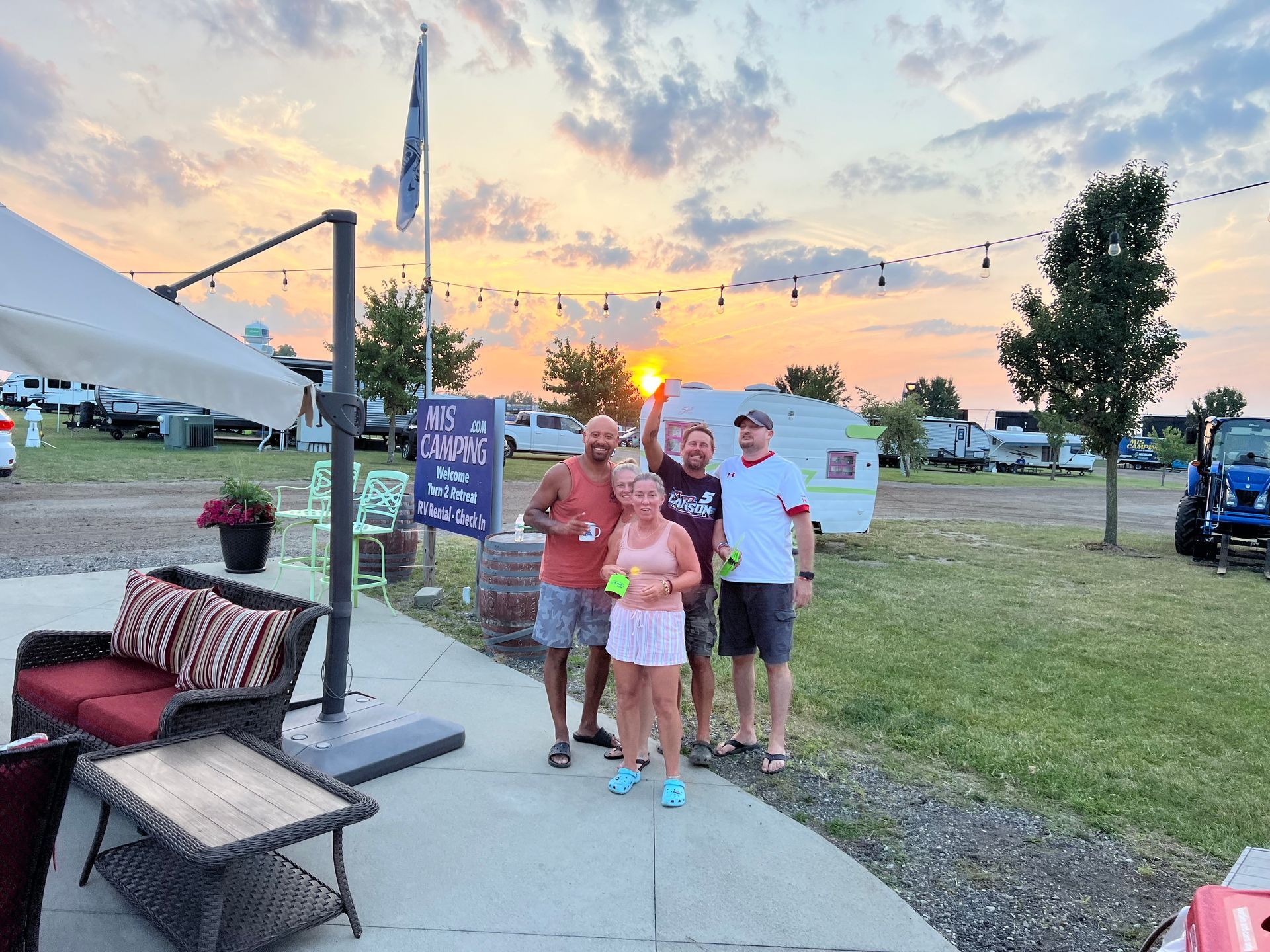 Four people cheering, holding drinks and pickleball balls, sunset in background.