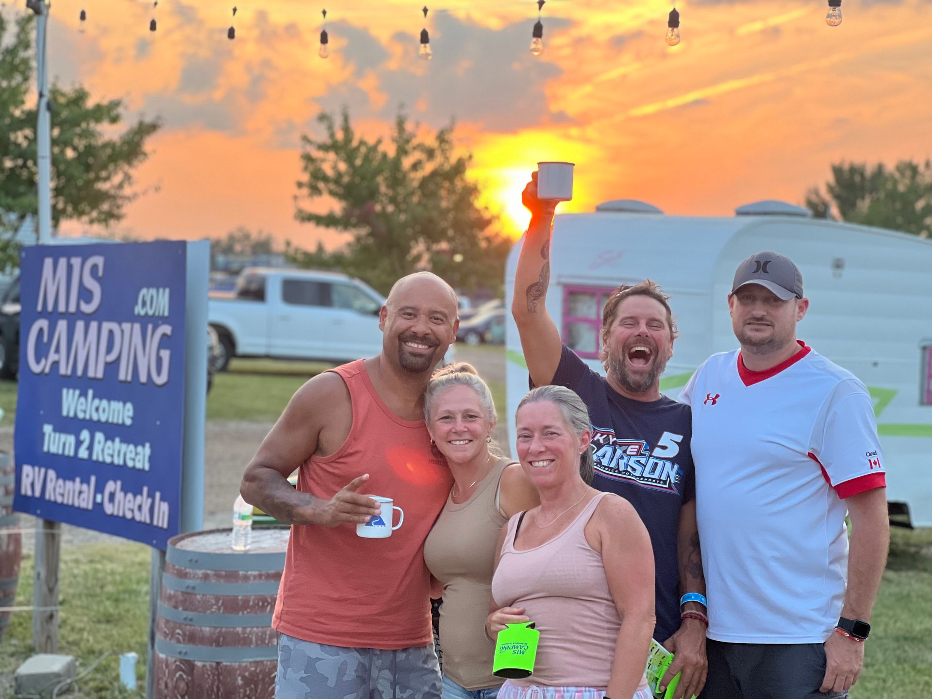 Group of five friends celebrating at a campsite with sunset. Holding mugs and smiling. Sign: 