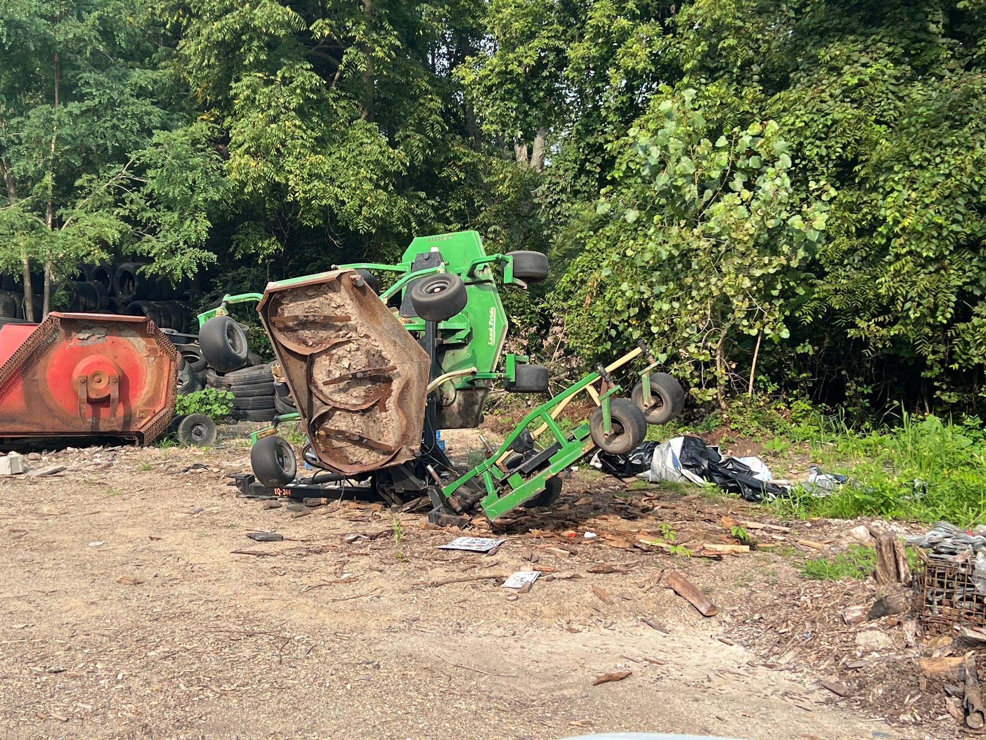 Green tractor overturned near trees and a red piece of farm equipment.
