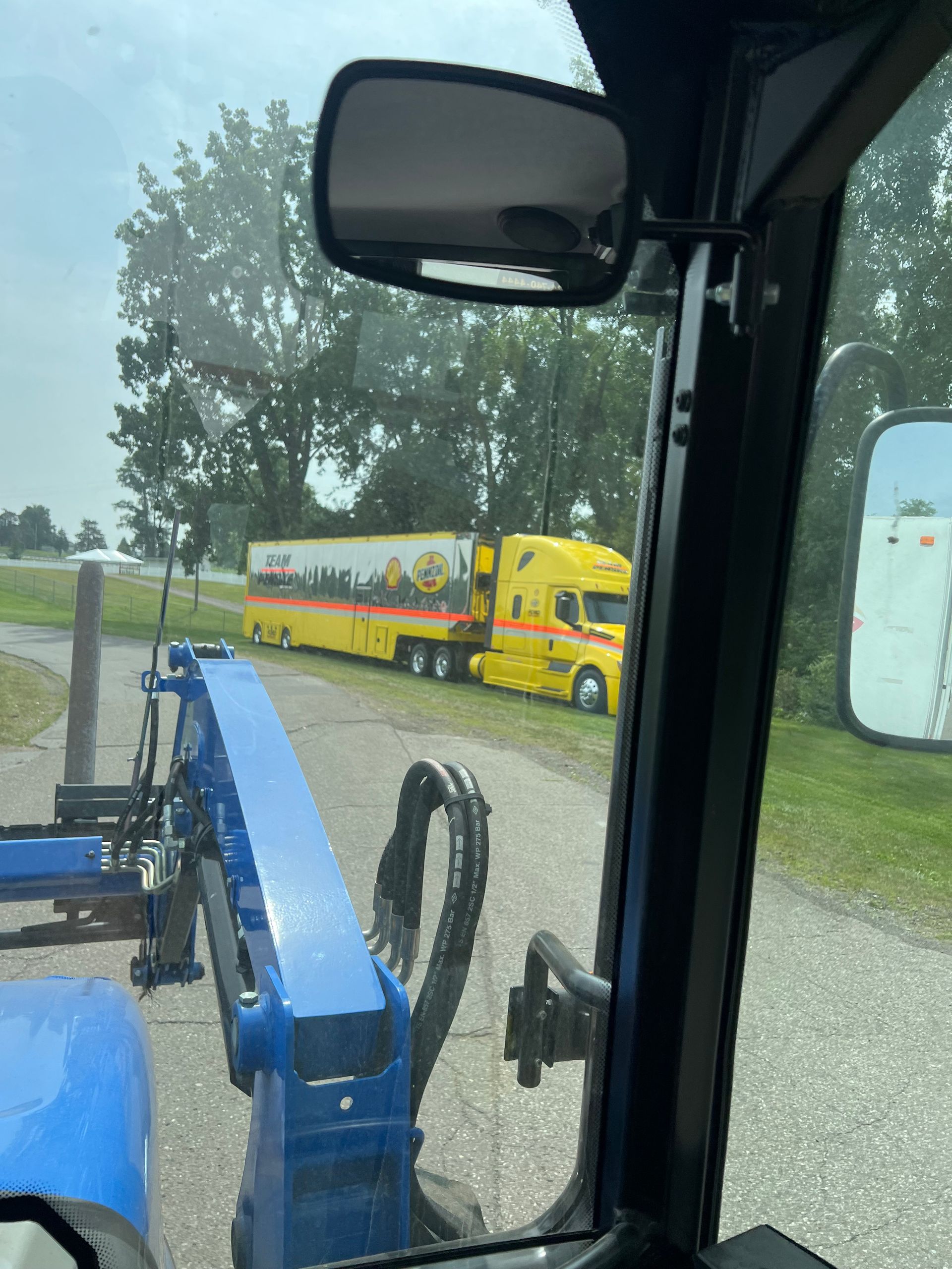 View from a tractor cab of a yellow semi-truck with trailer, parked on a gravel road next to trees.