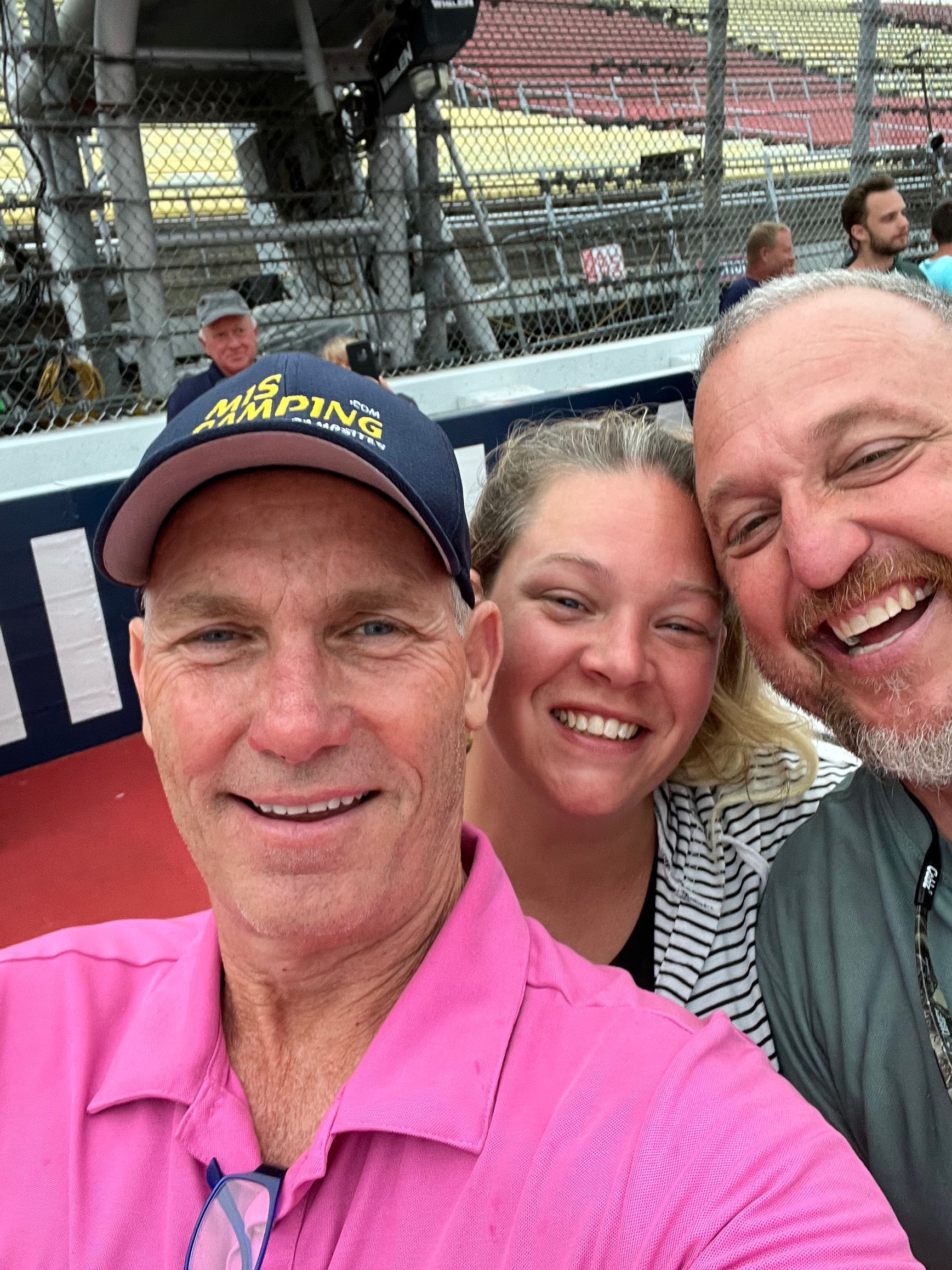 Three people smiling for a selfie at a racetrack; man in pink shirt, woman in striped shirt, man with beard.