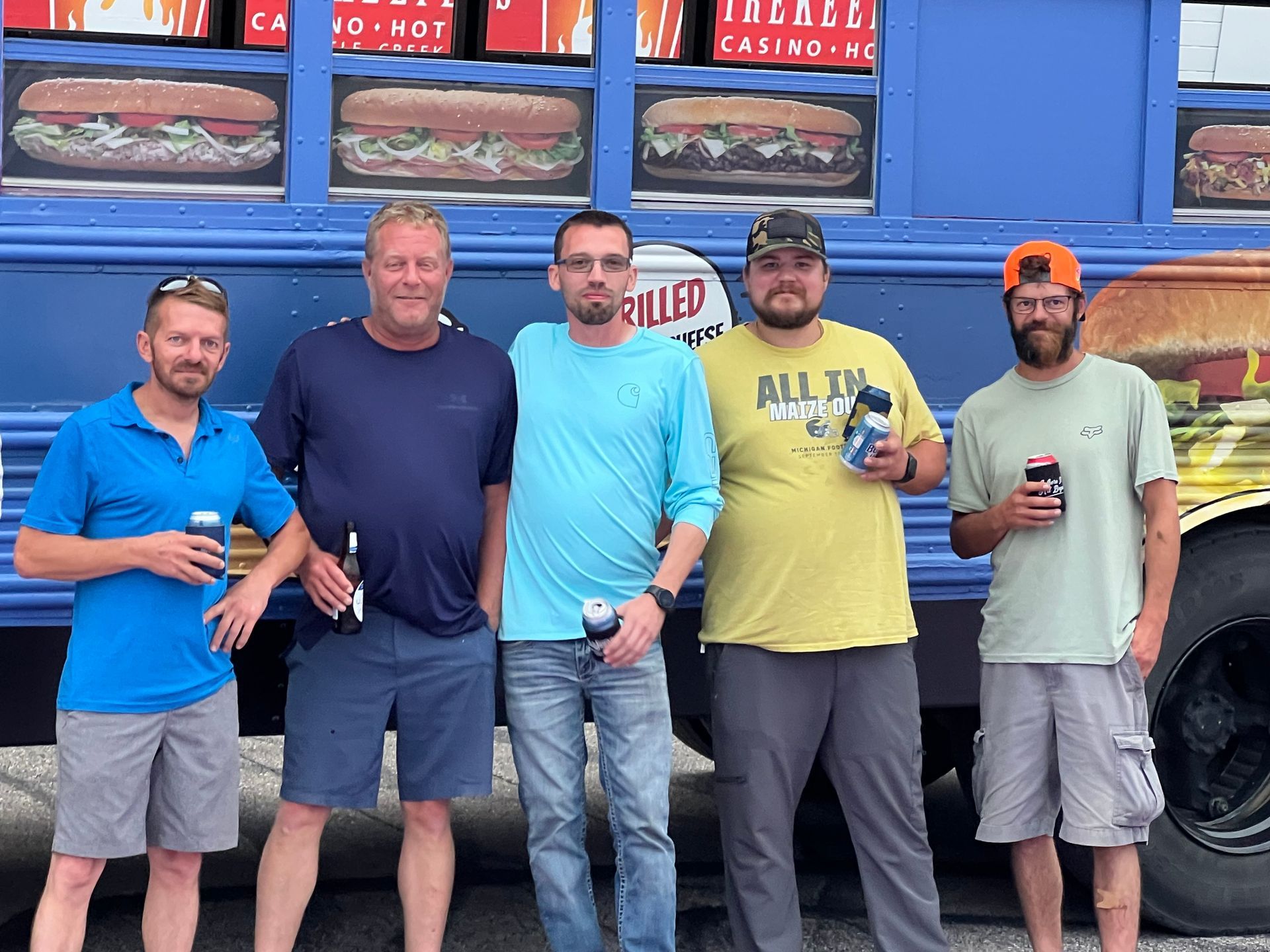 Five men pose in front of a blue food truck with sandwich images; they hold drinks and smile.