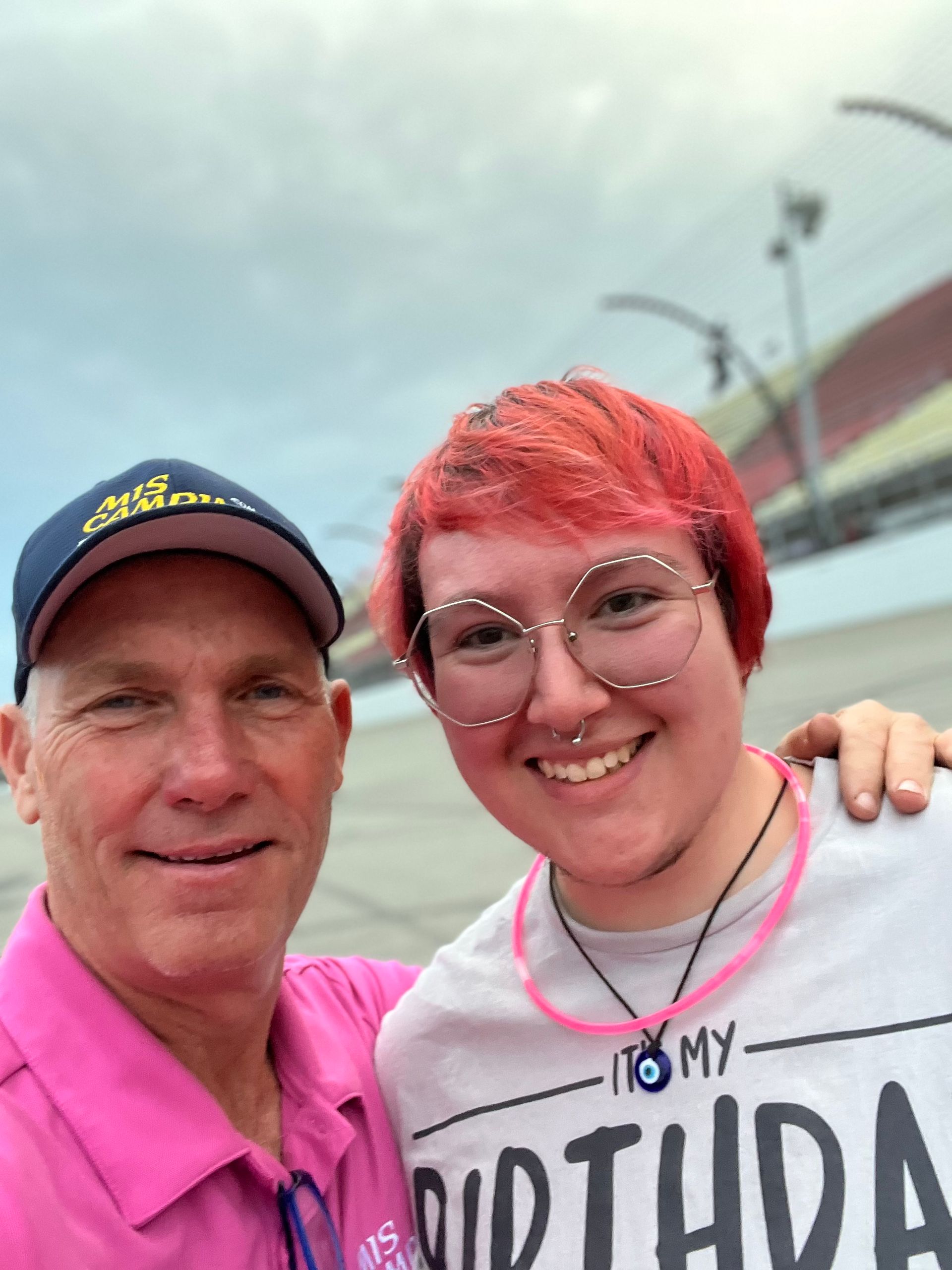 Man in pink shirt and cap smiles with person with red hair, at racetrack.