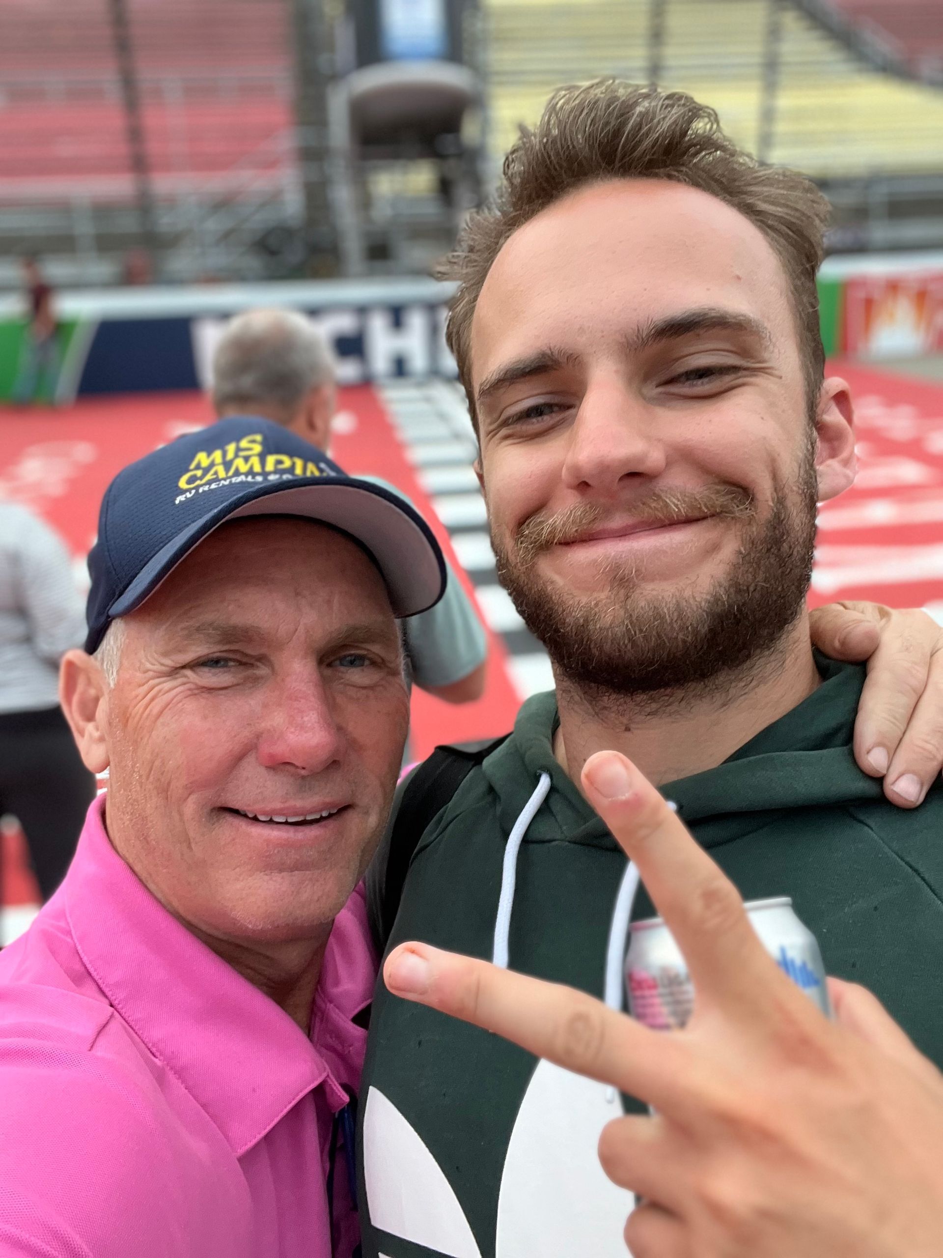 Two men smiling for a selfie; one in a pink shirt and hat, the other in a green hoodie, holding up a peace sign.