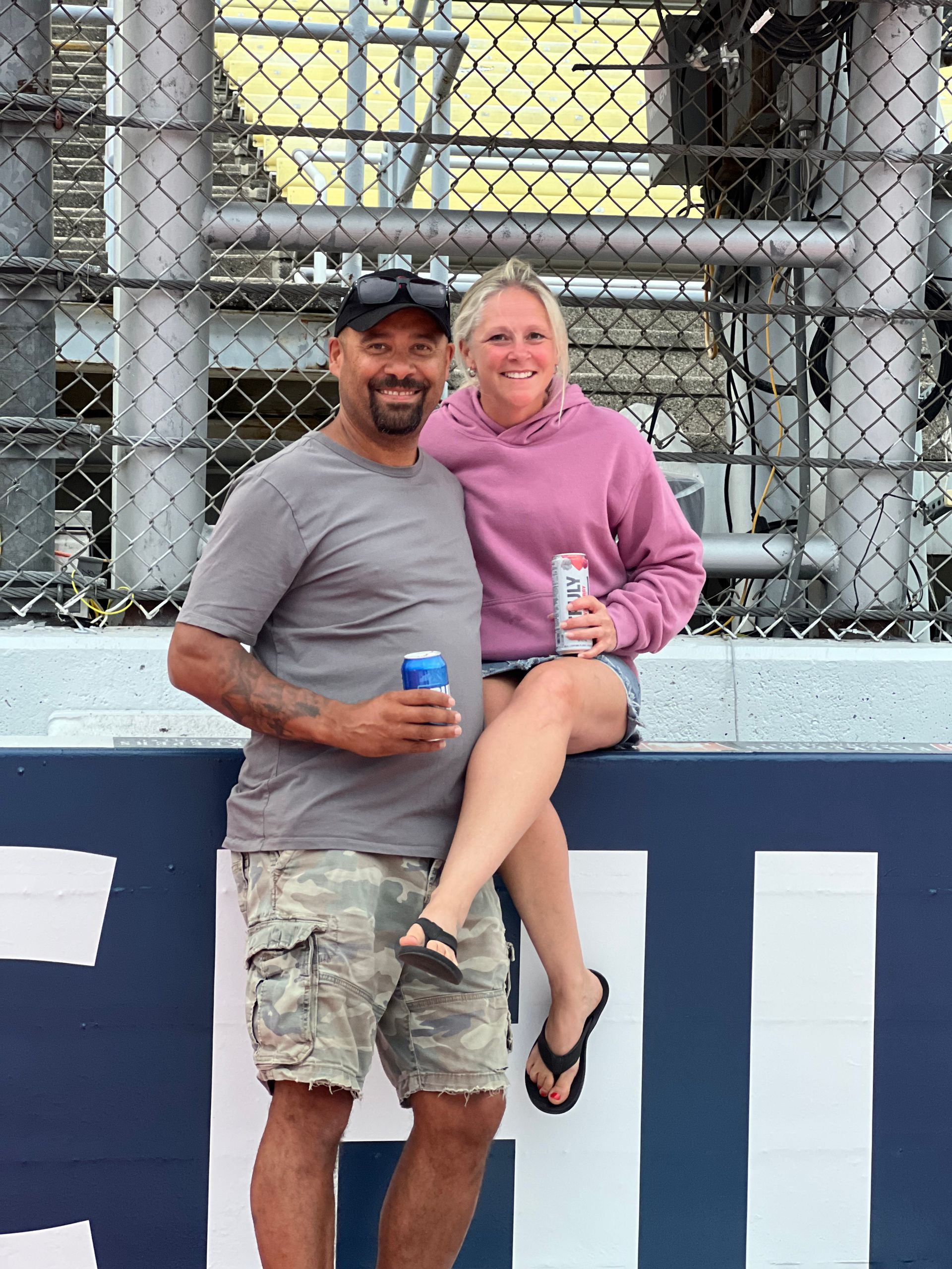 Couple smiling and holding drinks near a fence, man in camo shorts, woman in pink hoodie, at a racetrack.