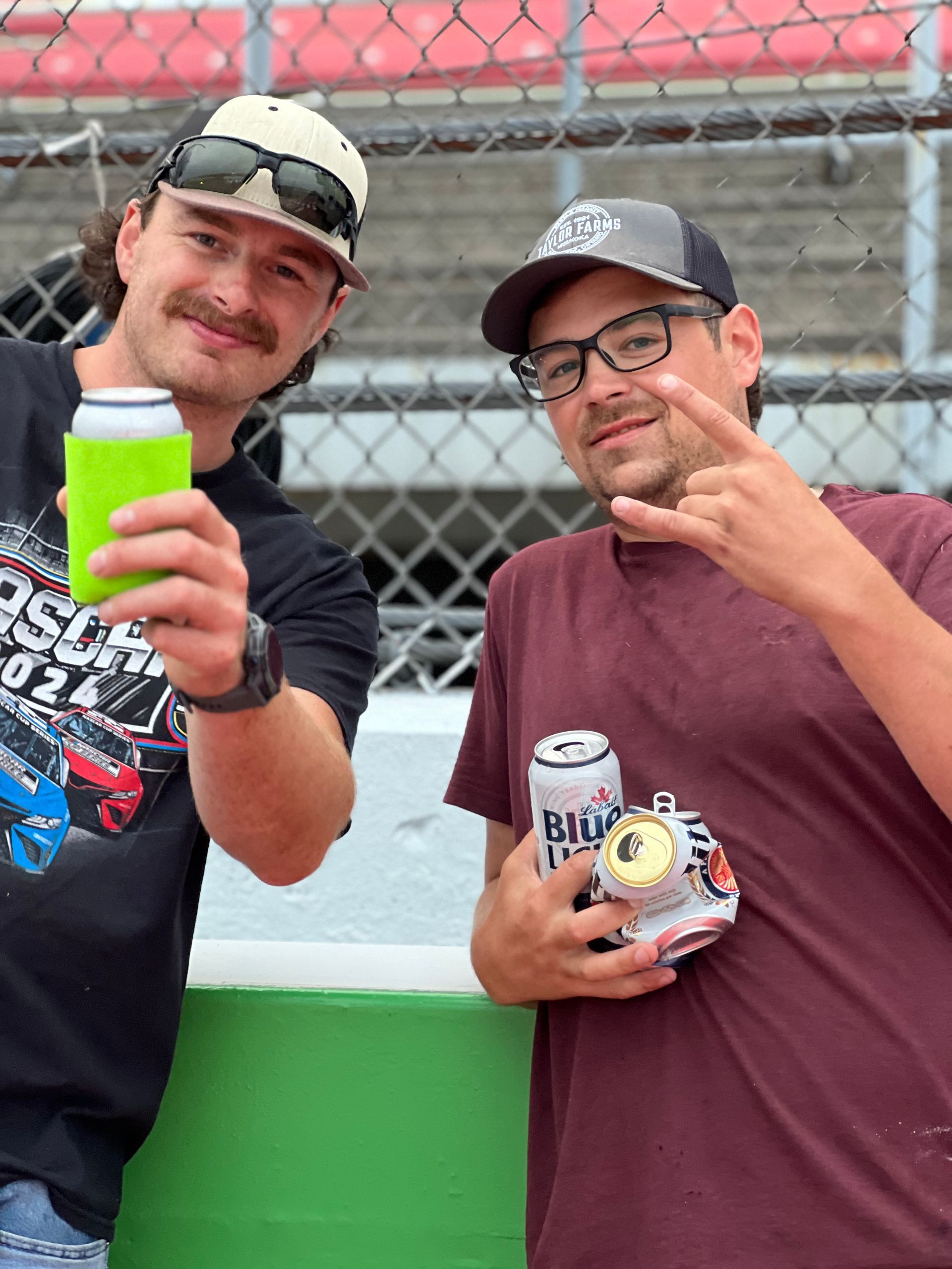 Two men at a racetrack, holding drinks. One man shows a rock on hand gesture.