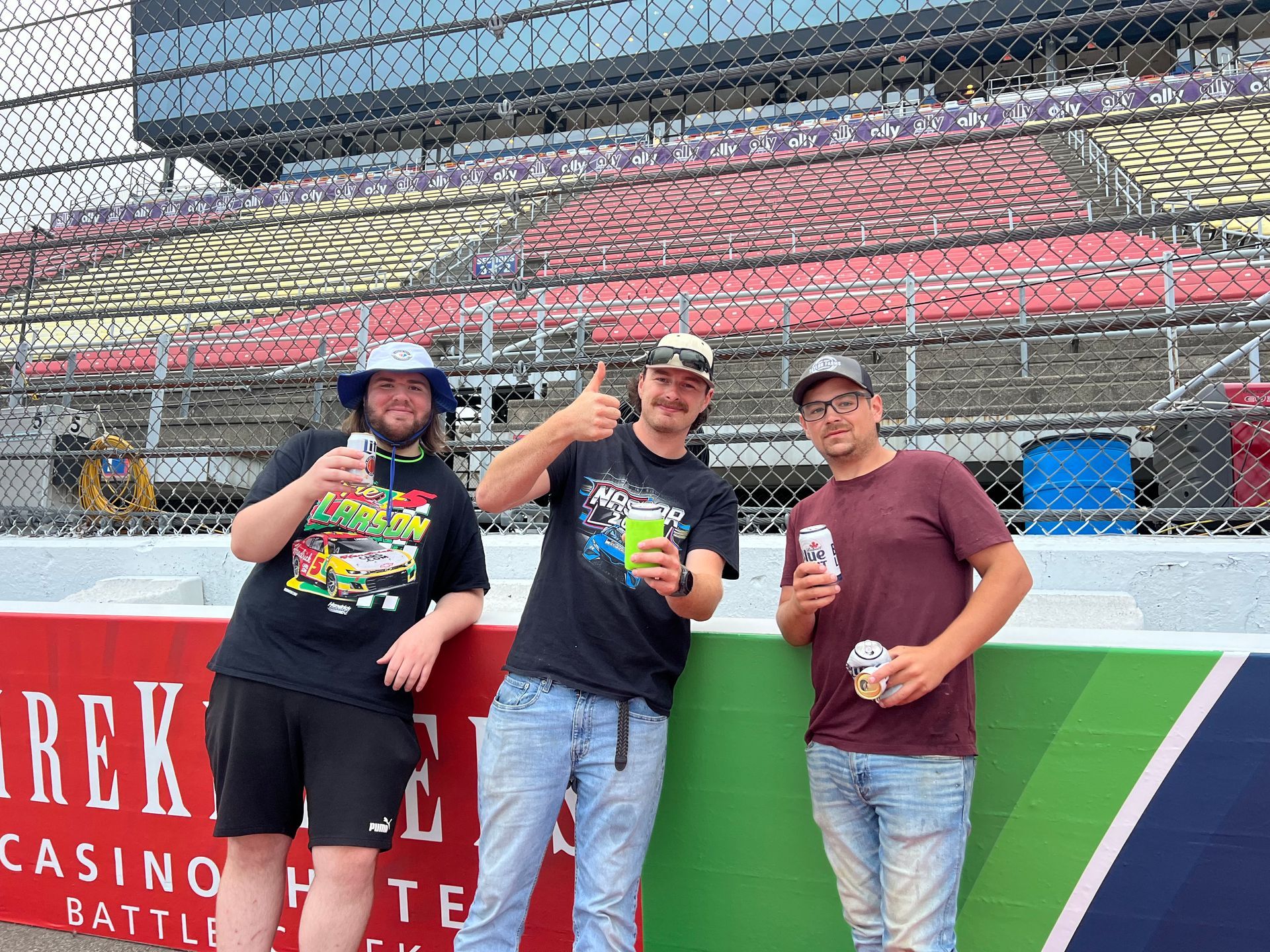 Three people holding beverages, posing near racetrack barriers; one gives a thumbs-up.