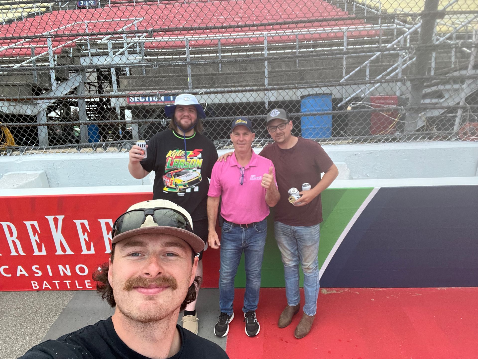 Four men pose at a racetrack; one takes a selfie..