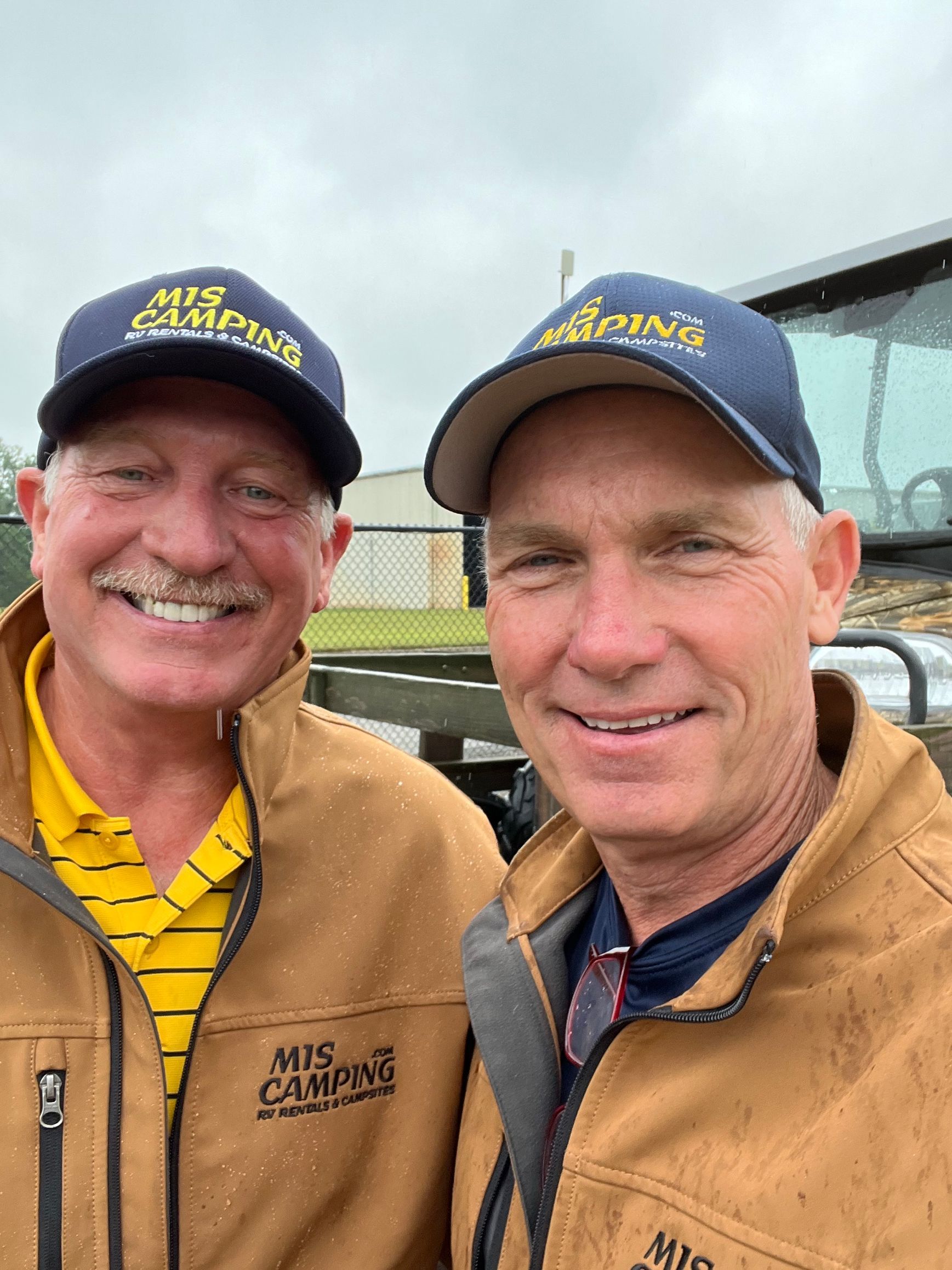 Two men in matching hats and jackets, smiling, near construction equipment.