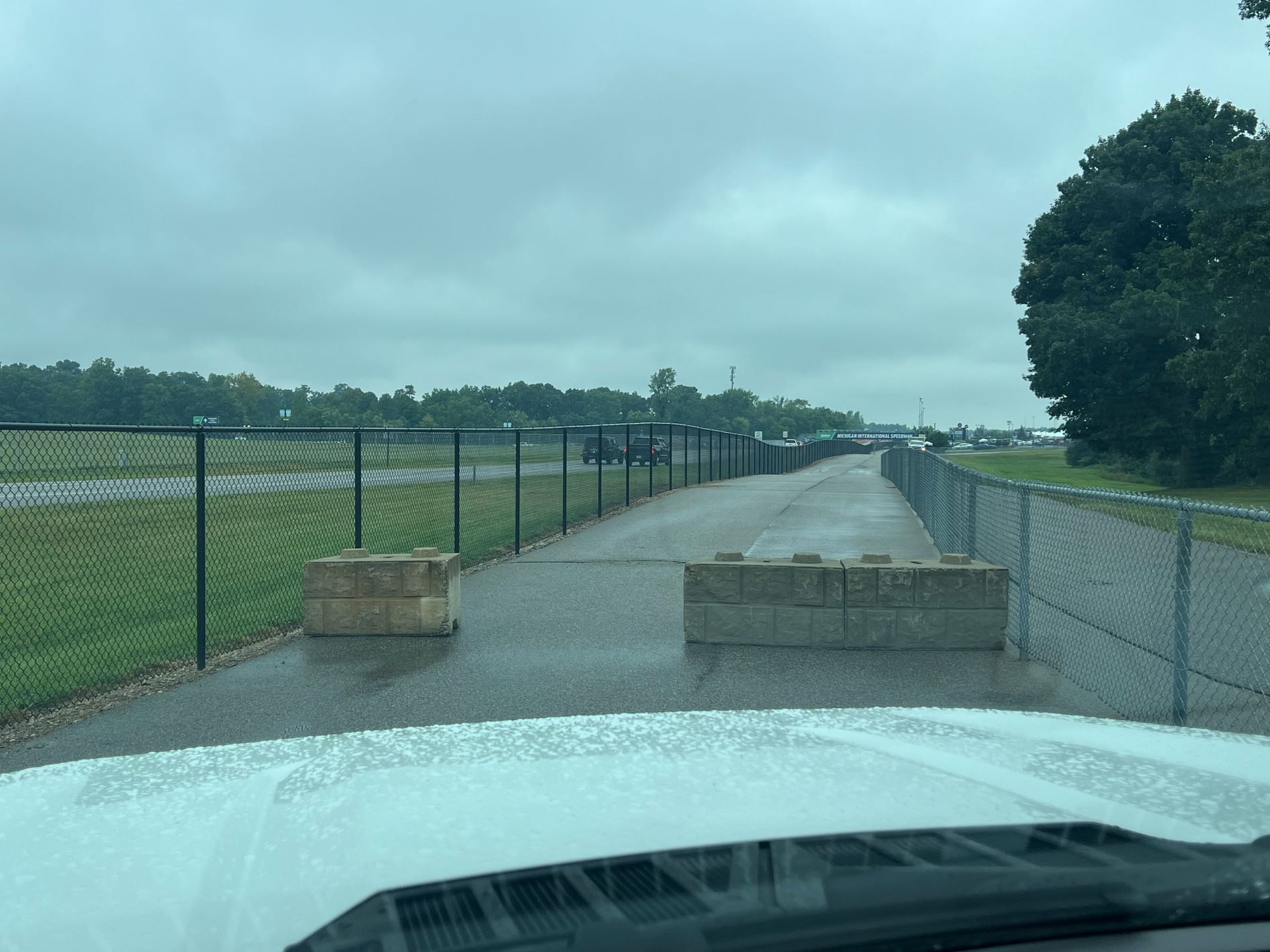 View from a vehicle's windshield; wet road blocked by concrete barriers, flanked by chain-link fences and a green field.