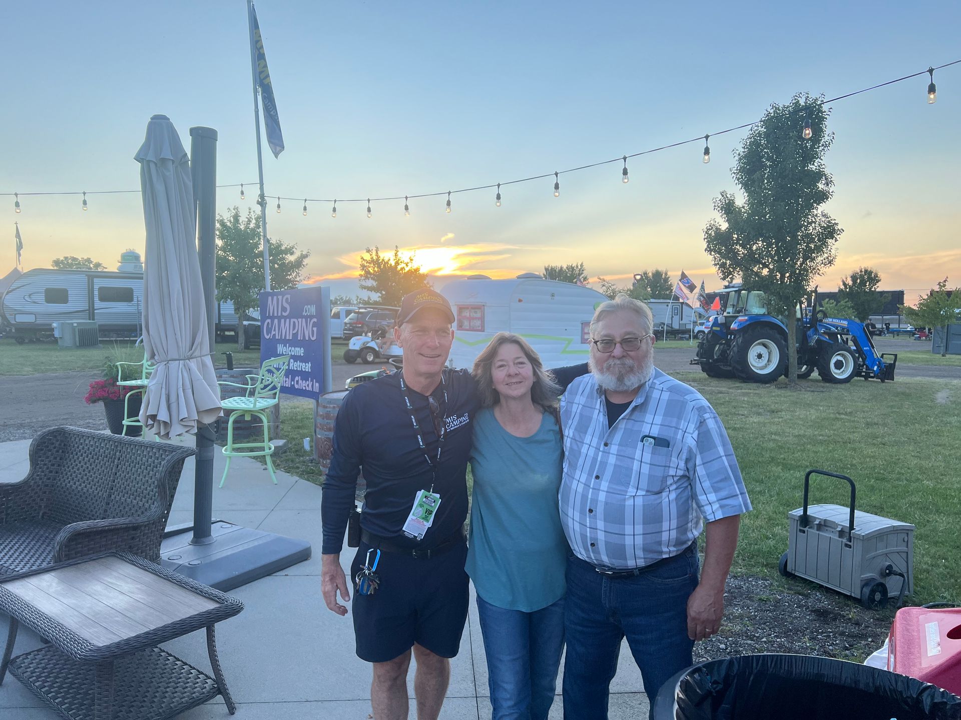 Three people smiling outdoors near picnic tables and RVs at sunset.