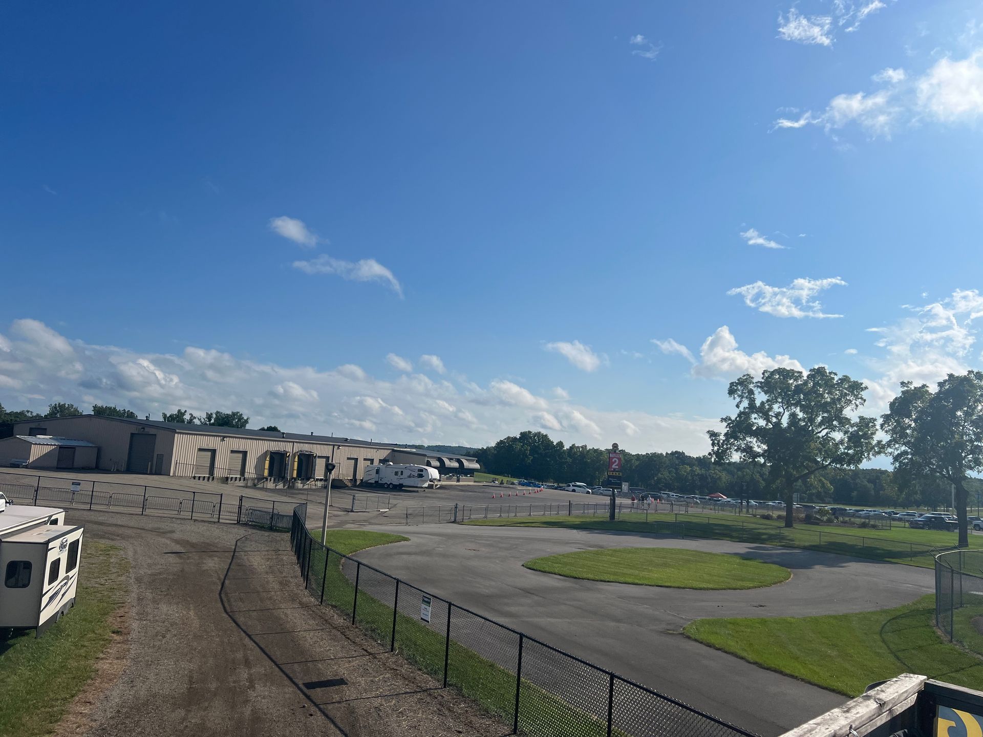 Open area with buildings, parked vehicles, and trees under a partly cloudy blue sky.