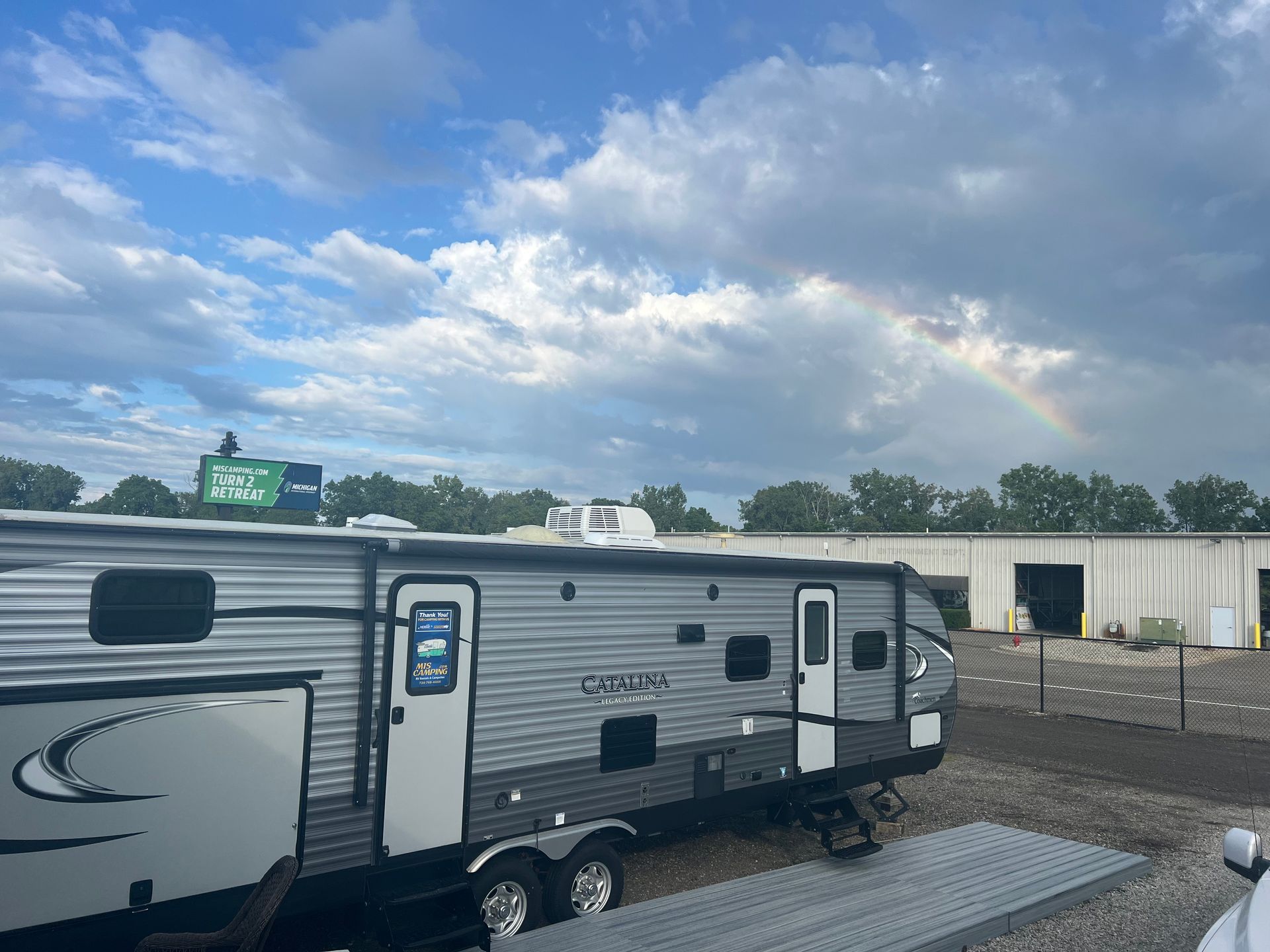 RV trailer parked outside under a partly cloudy sky with a rainbow visible.