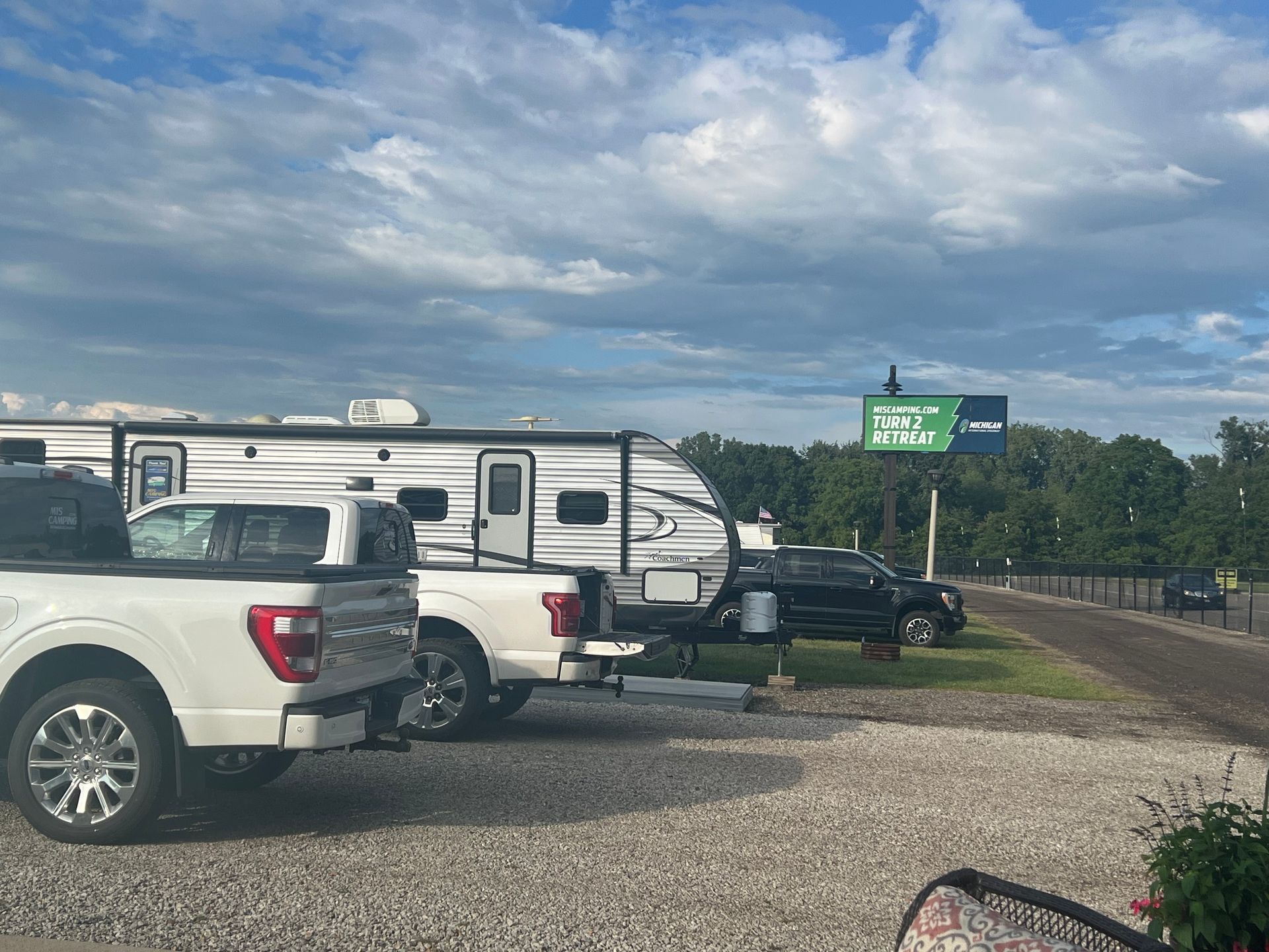 RVs and trucks parked on gravel at campsite under cloudy sky, with road sign in the distance.