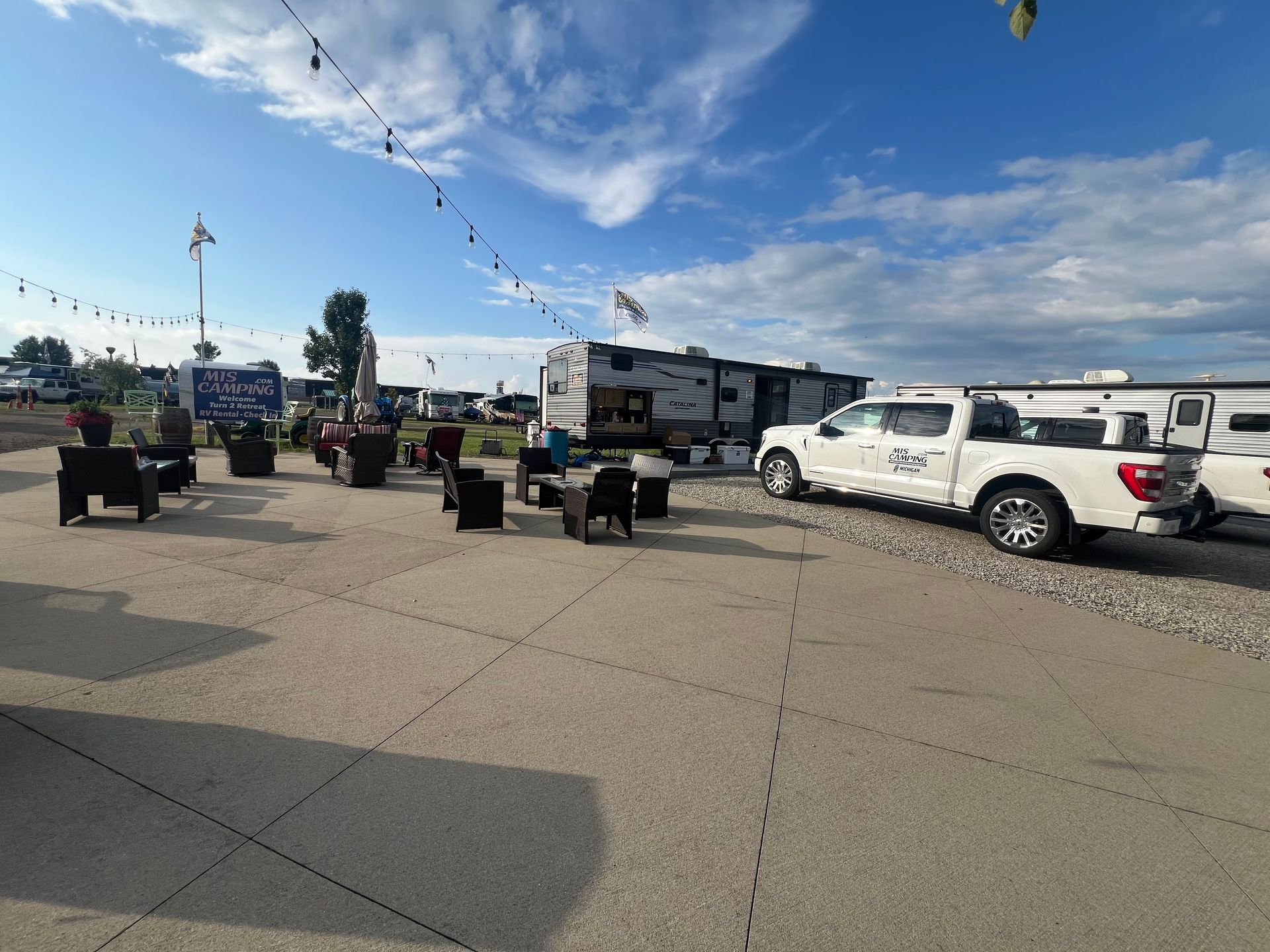 Campground with RVs, a white pickup truck, and chairs on a gravel lot under a partly cloudy sky.