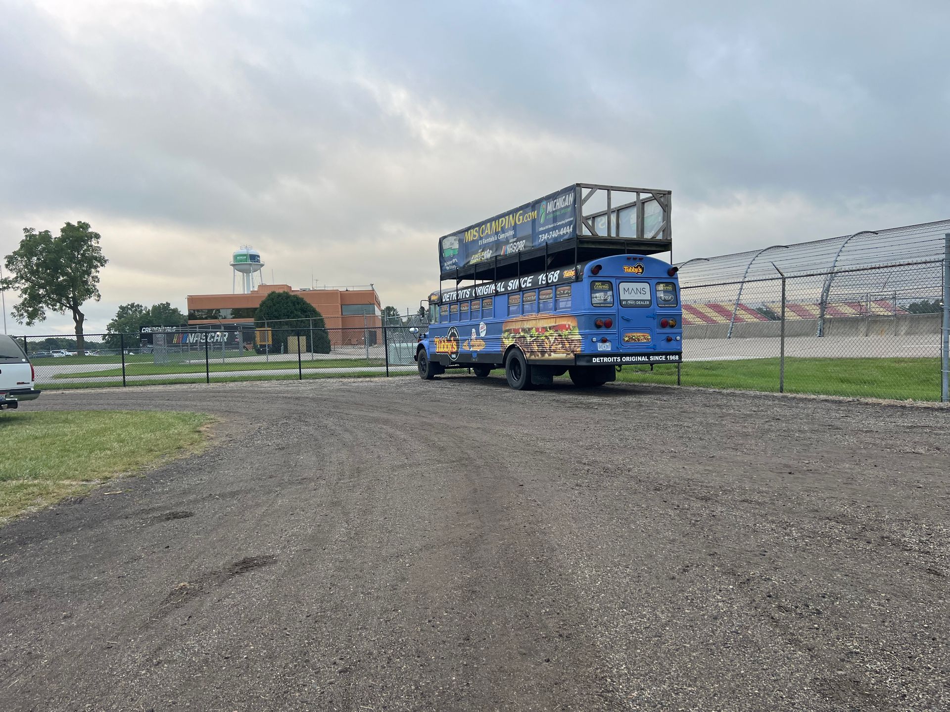 Blue bus with a double-decker structure, parked on gravel, with a building and cloudy sky in the background.