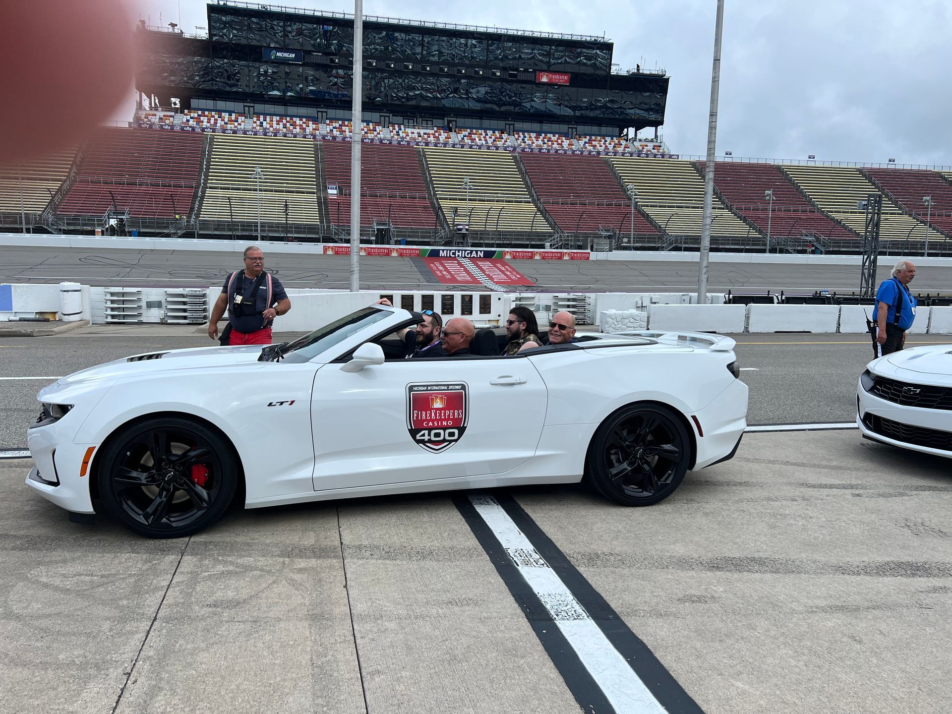 White convertible car on racetrack, people inside. Spectator seating in background.