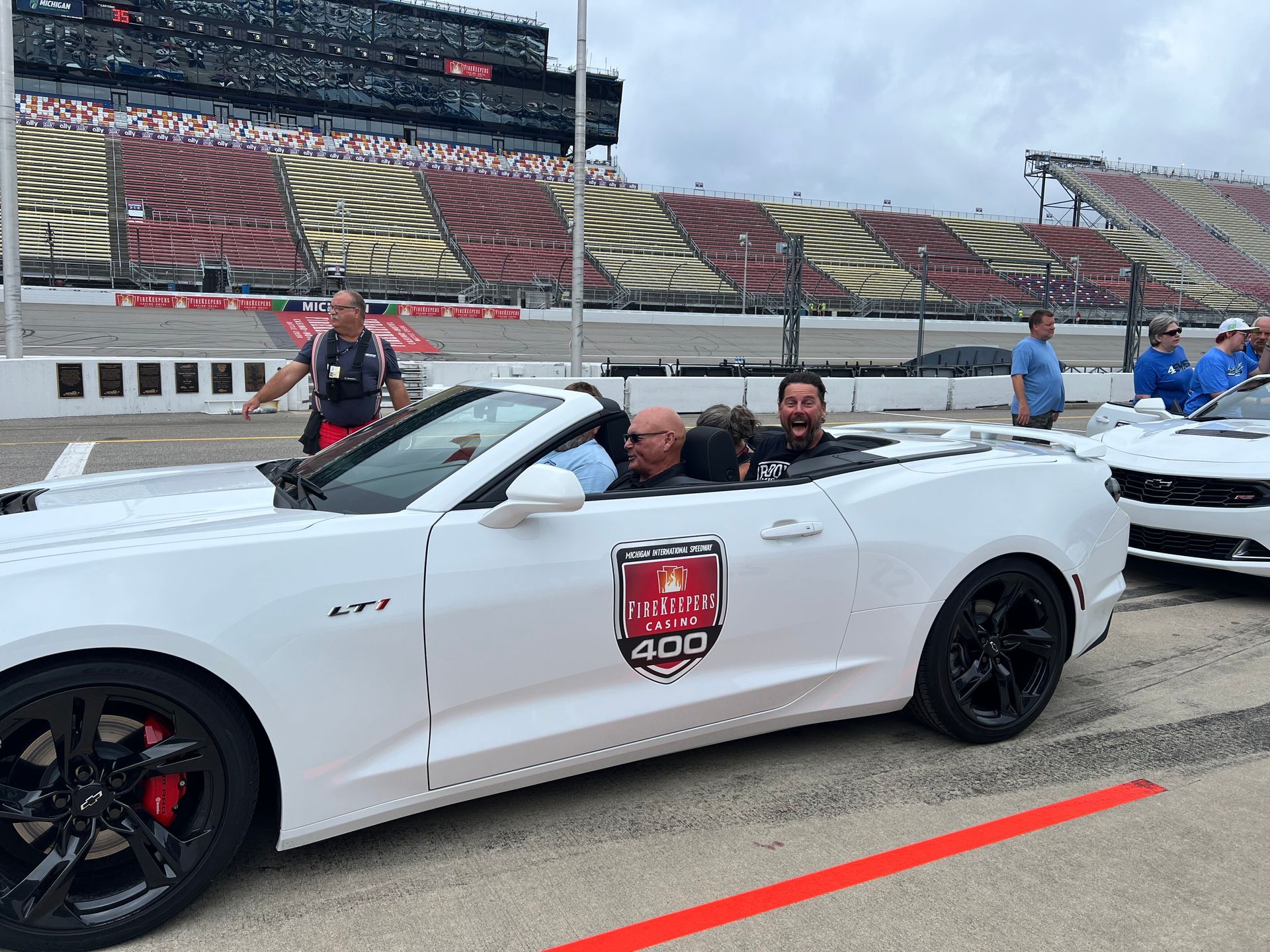 White convertible car with passengers on racetrack, large stadium in background.