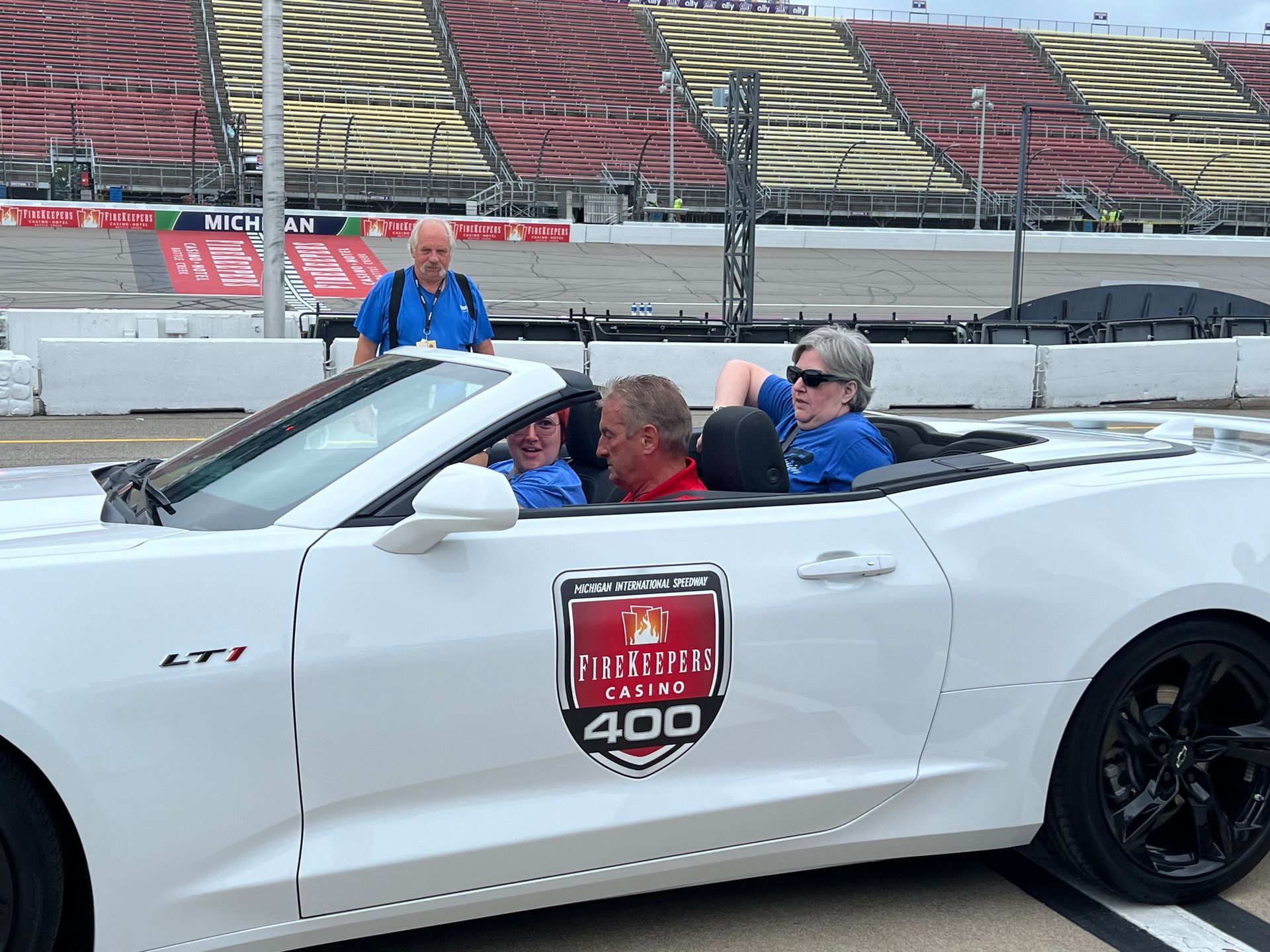 People in a white convertible car at a racetrack, a man stands in background.
