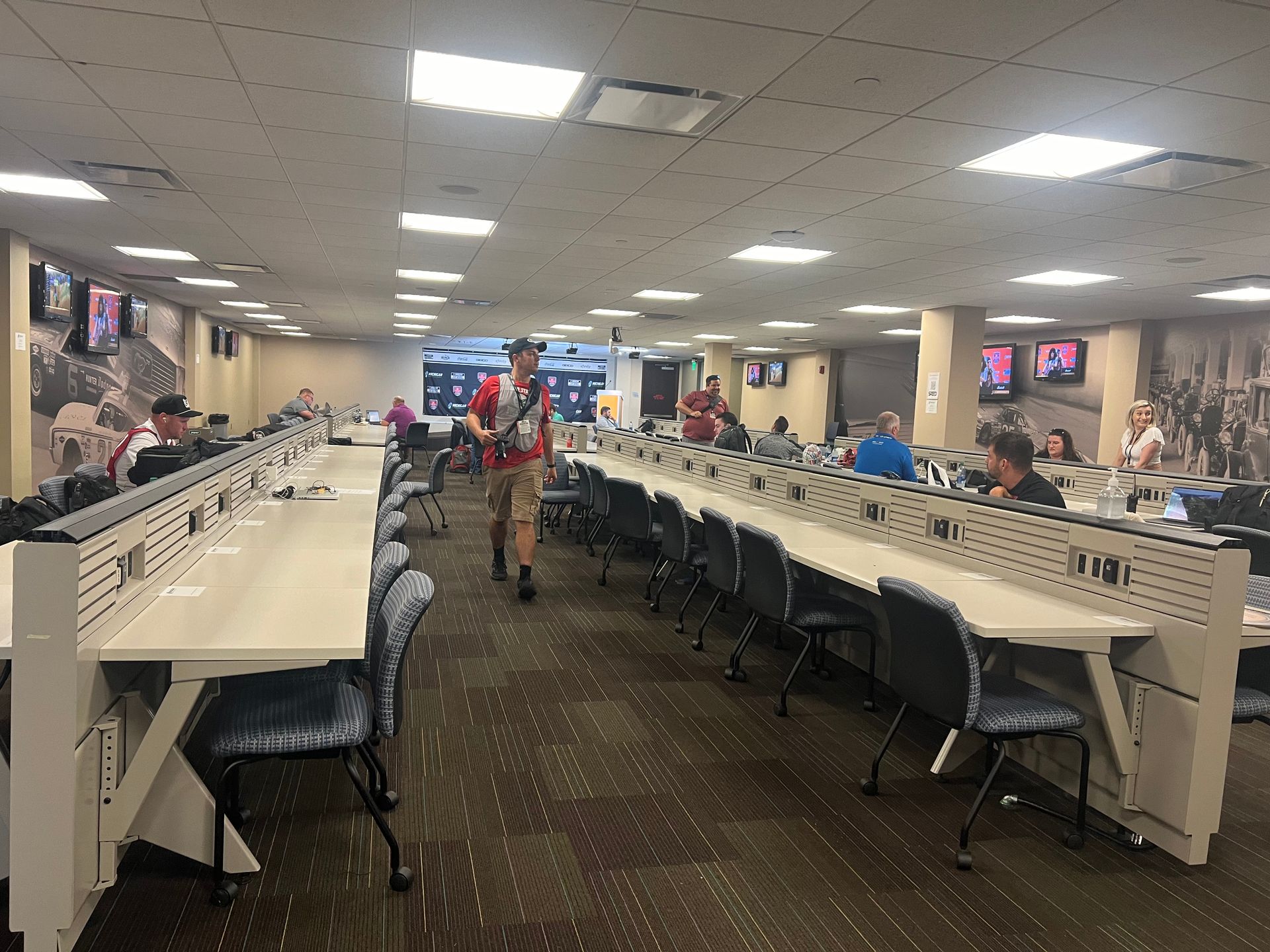 People in a large room with long tables, likely a betting area. TV screens on walls. One person walks through the space.