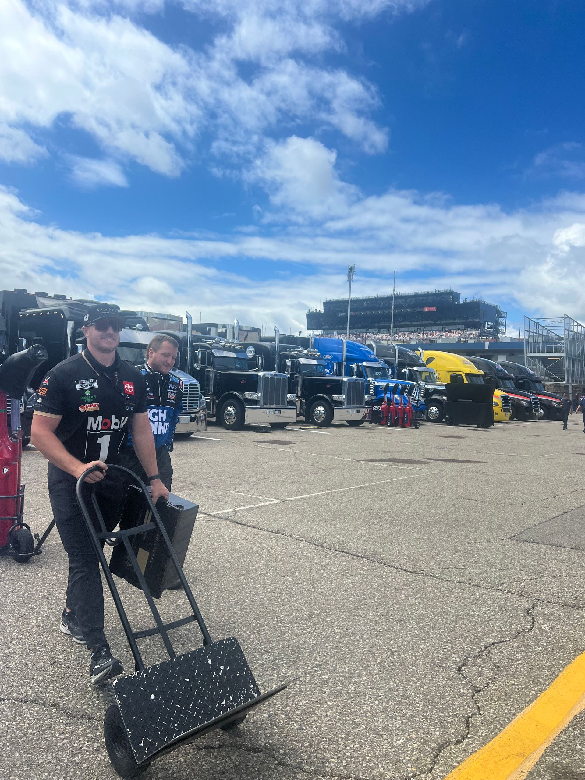 Man pushing a cart near semi-trucks at a racetrack. Blue sky.