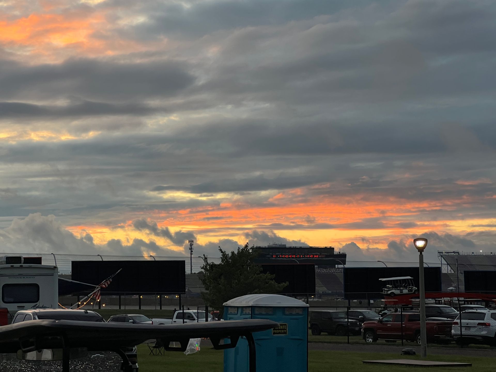 Sunset over a drive-in theater with dark screens, parked cars, and a portable toilet.