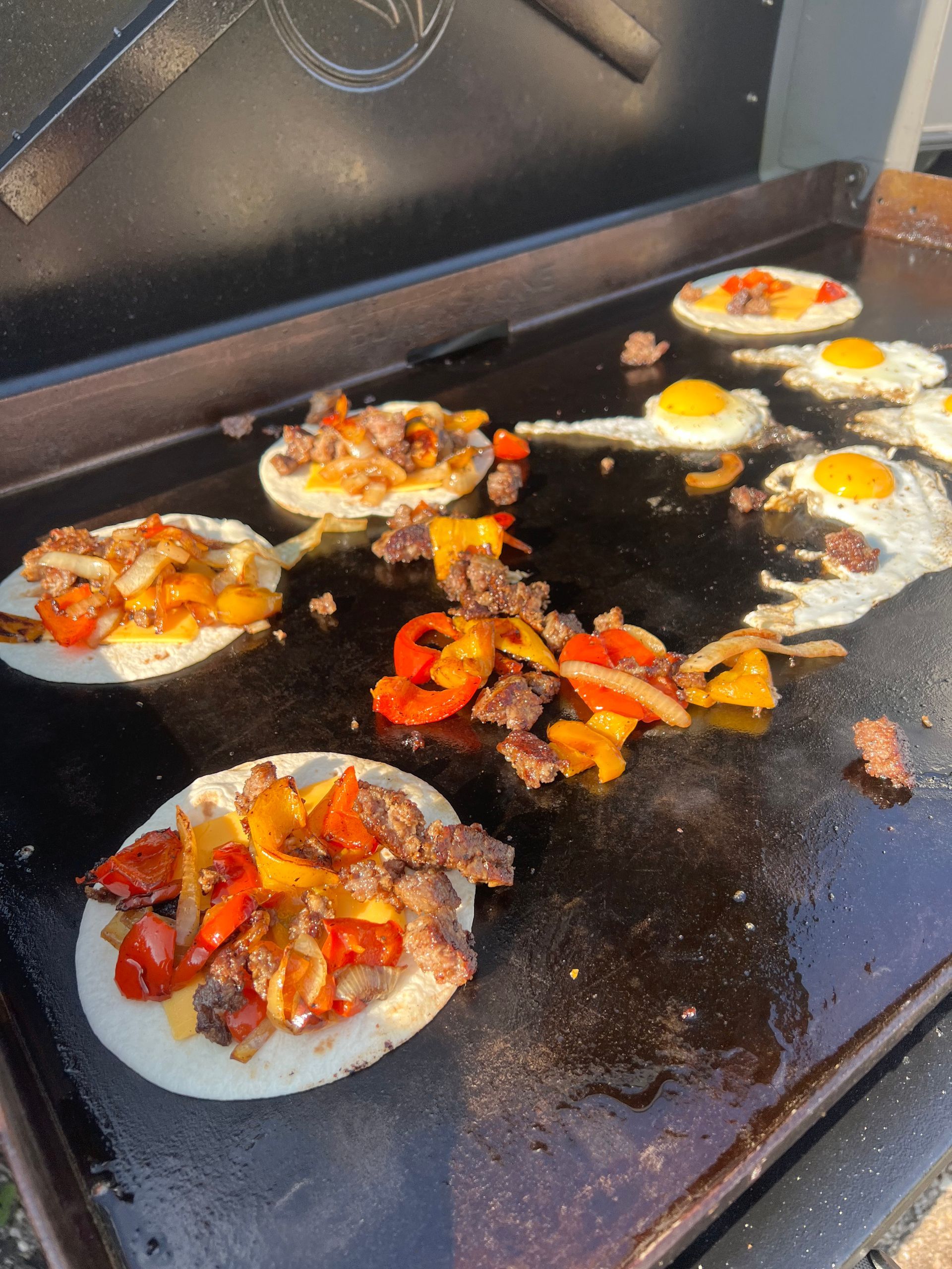 Flat top grill with breakfast tacos, fried eggs, and colorful peppers.
