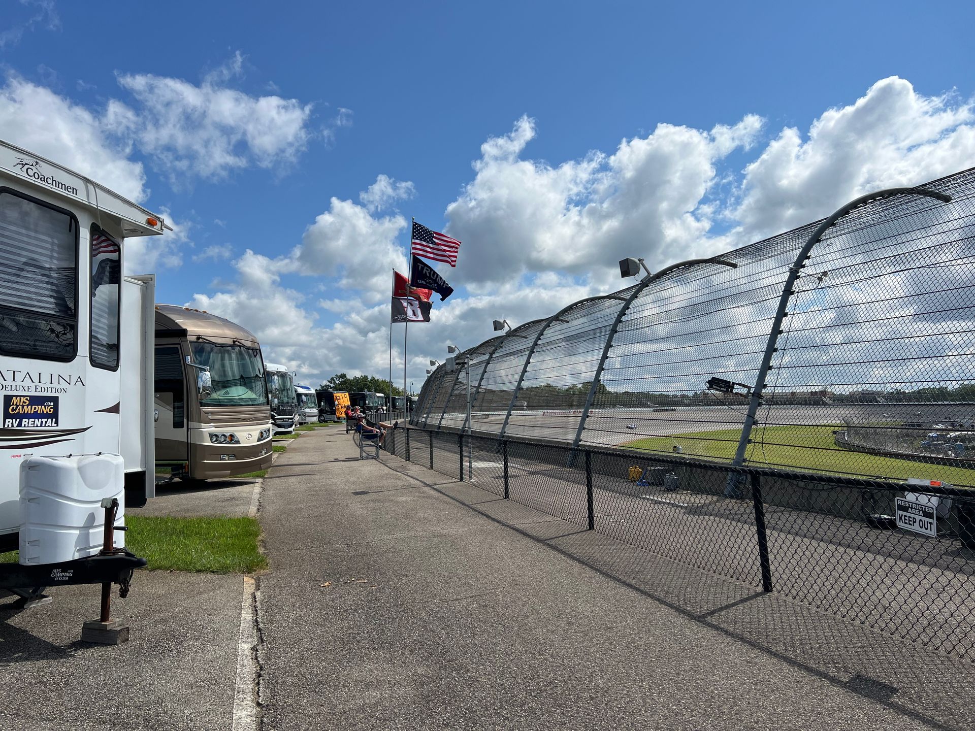 Campers line a paved walkway near a fence with flags under a partly cloudy sky.
