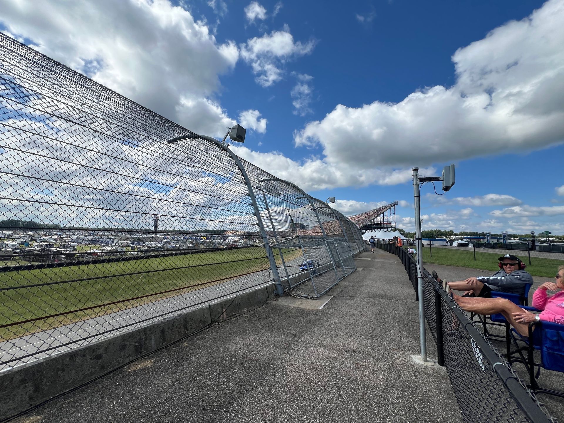 Race track fence with people watching under a blue sky with clouds.