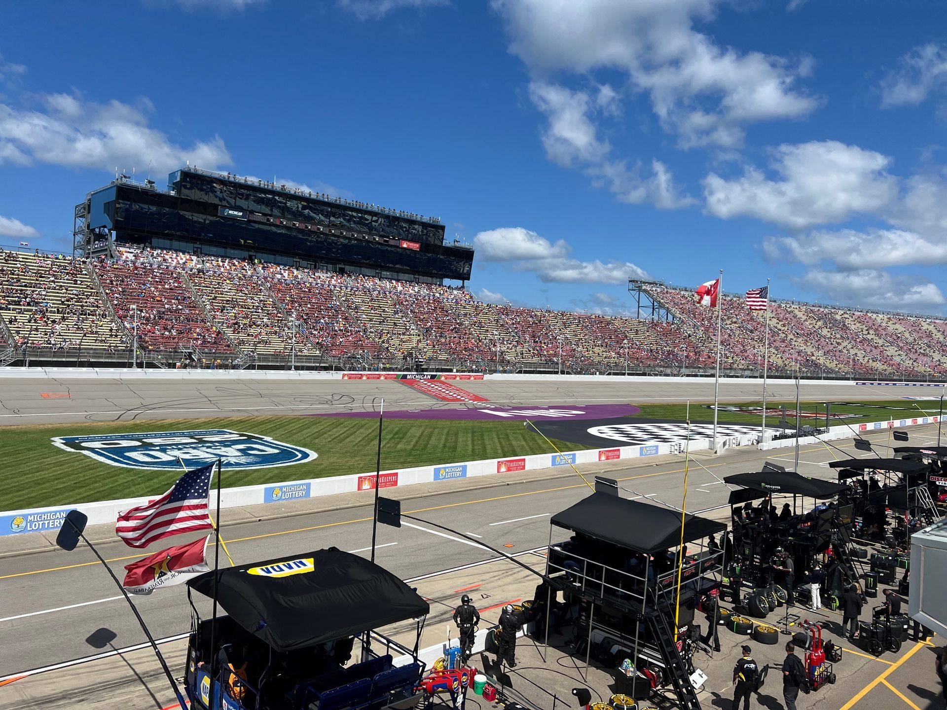 Race track with packed grandstands under a blue sky, pit crew area in foreground.