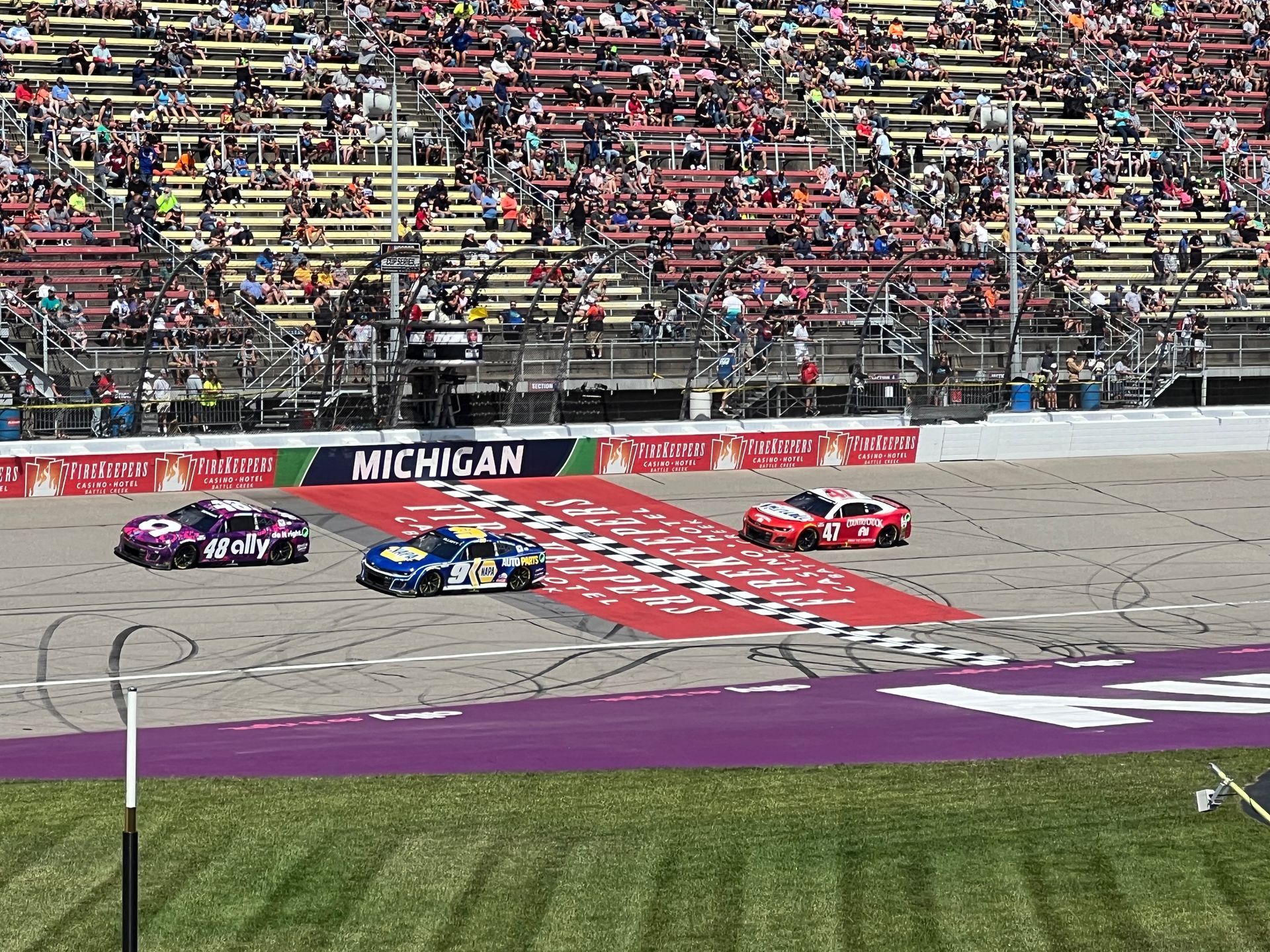 Race cars crossing finish line at Michigan International Speedway with crowded grandstands in background.