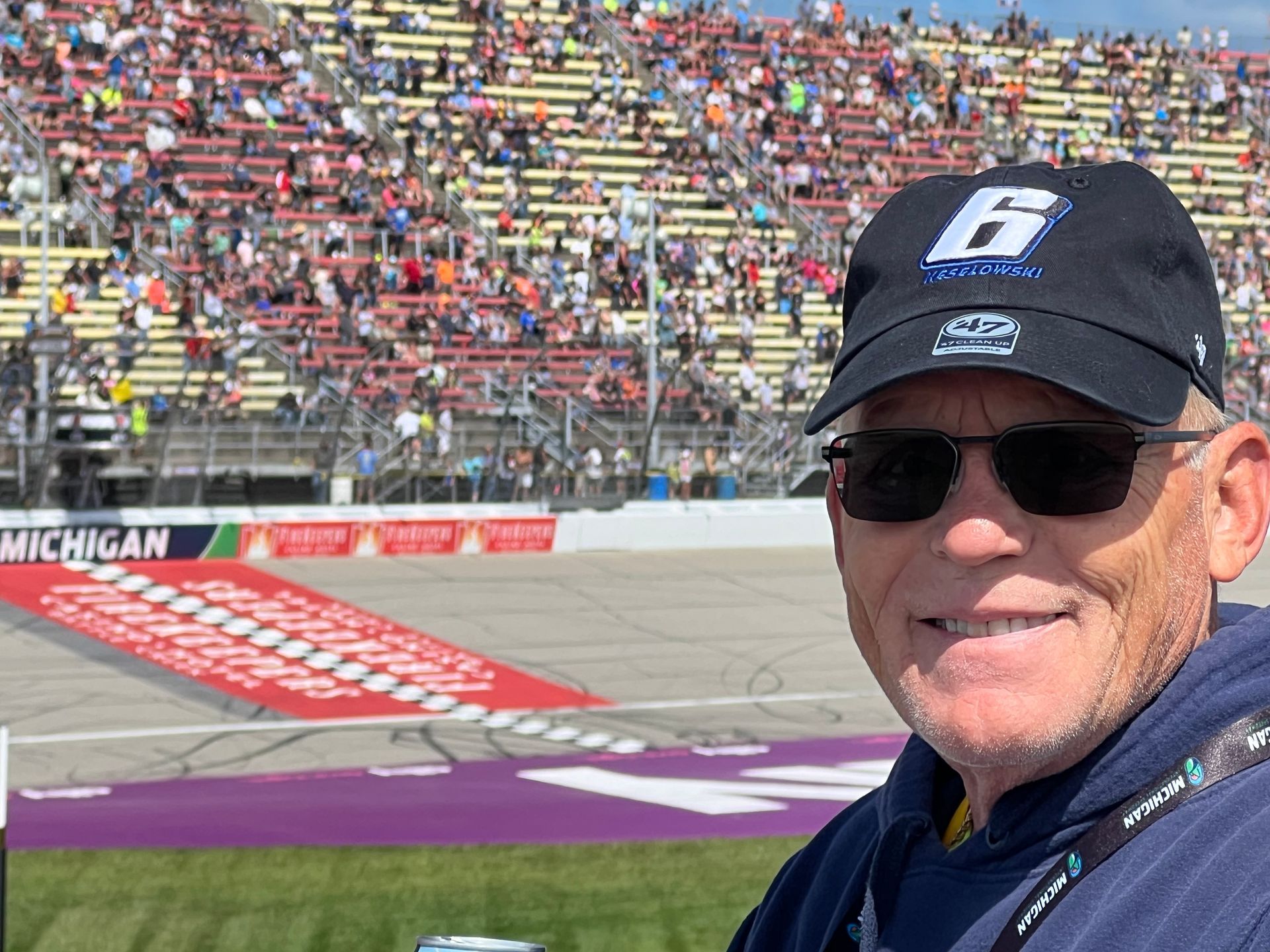 Man in sunglasses and hat smiles at camera; racetrack and crowd in background.