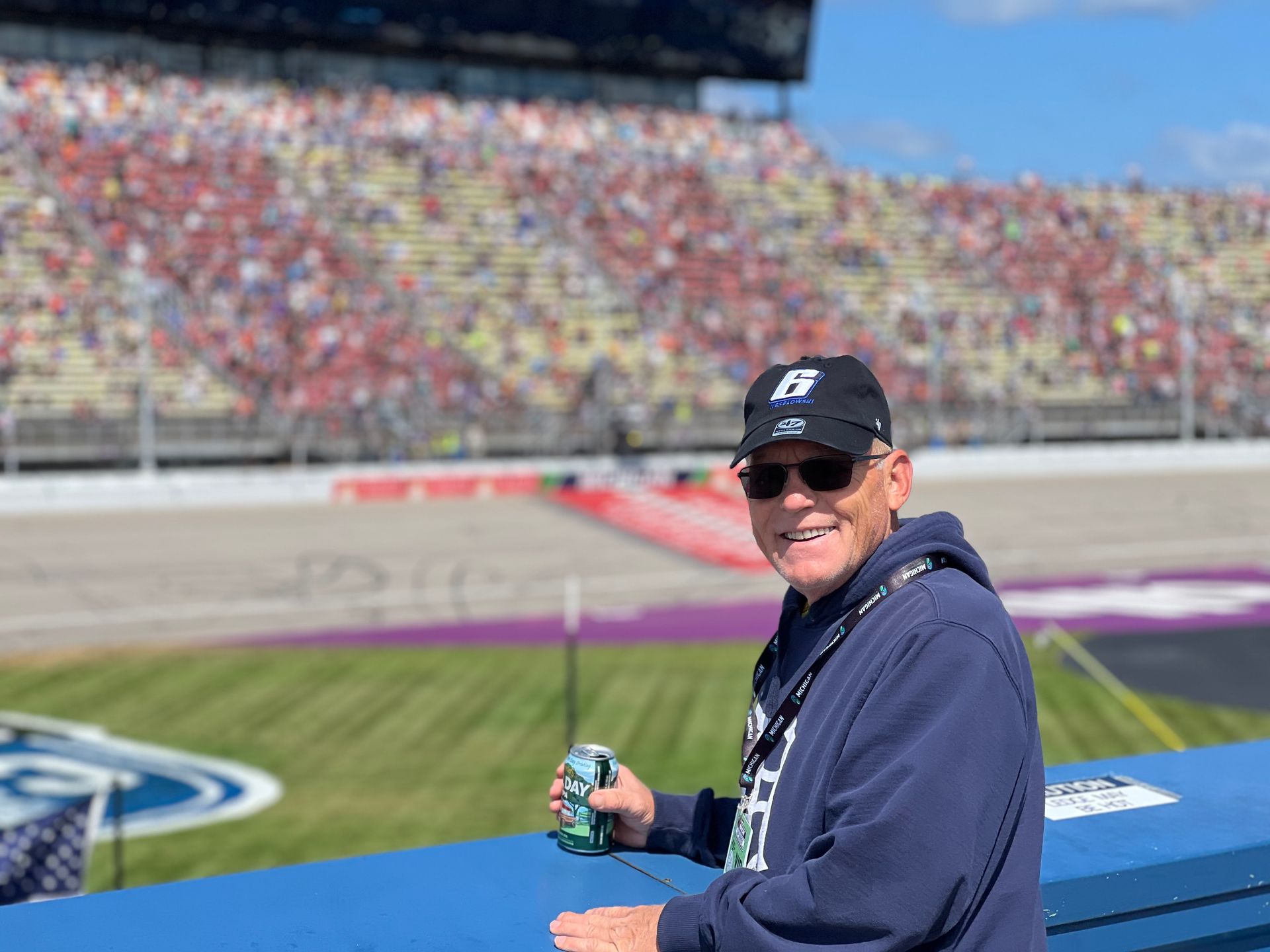 Man in a cap, smiling, holding a drink, at a racetrack with spectators in the background.