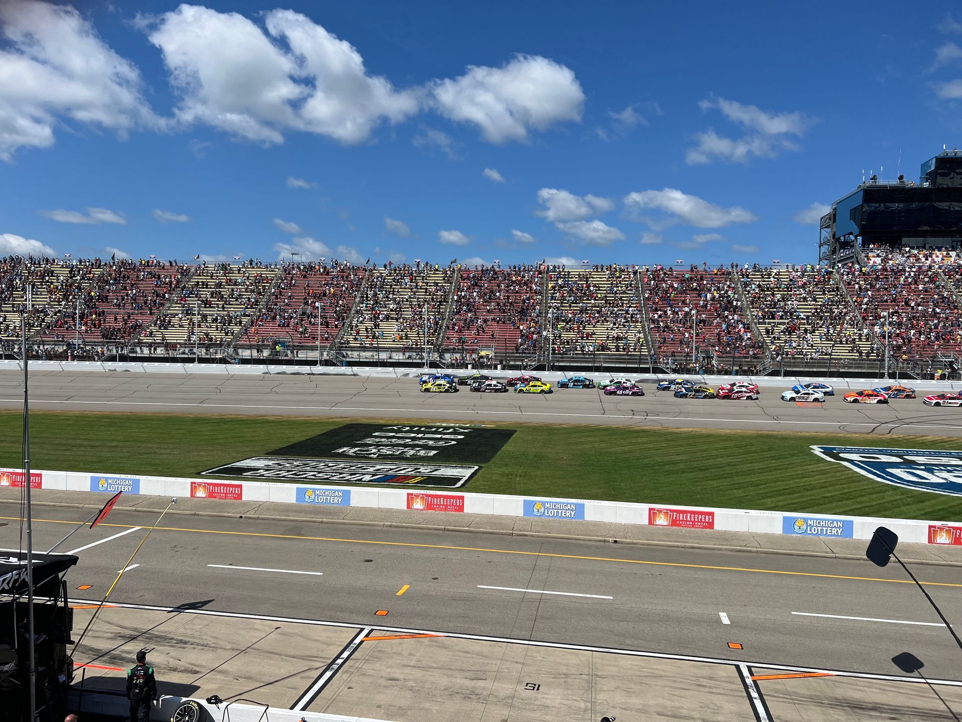 Race cars on a track, spectators in the stands, blue sky with clouds.