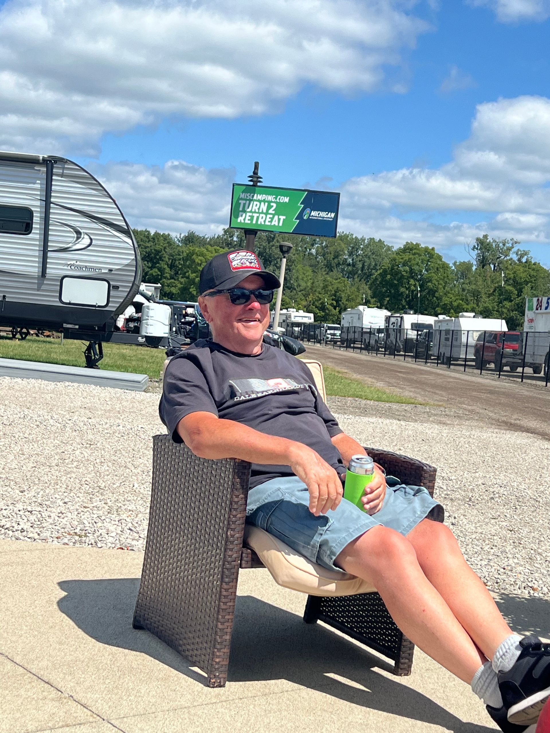 Man in sunglasses seated in chair outdoors, holding a drink. RVs and trees in background.