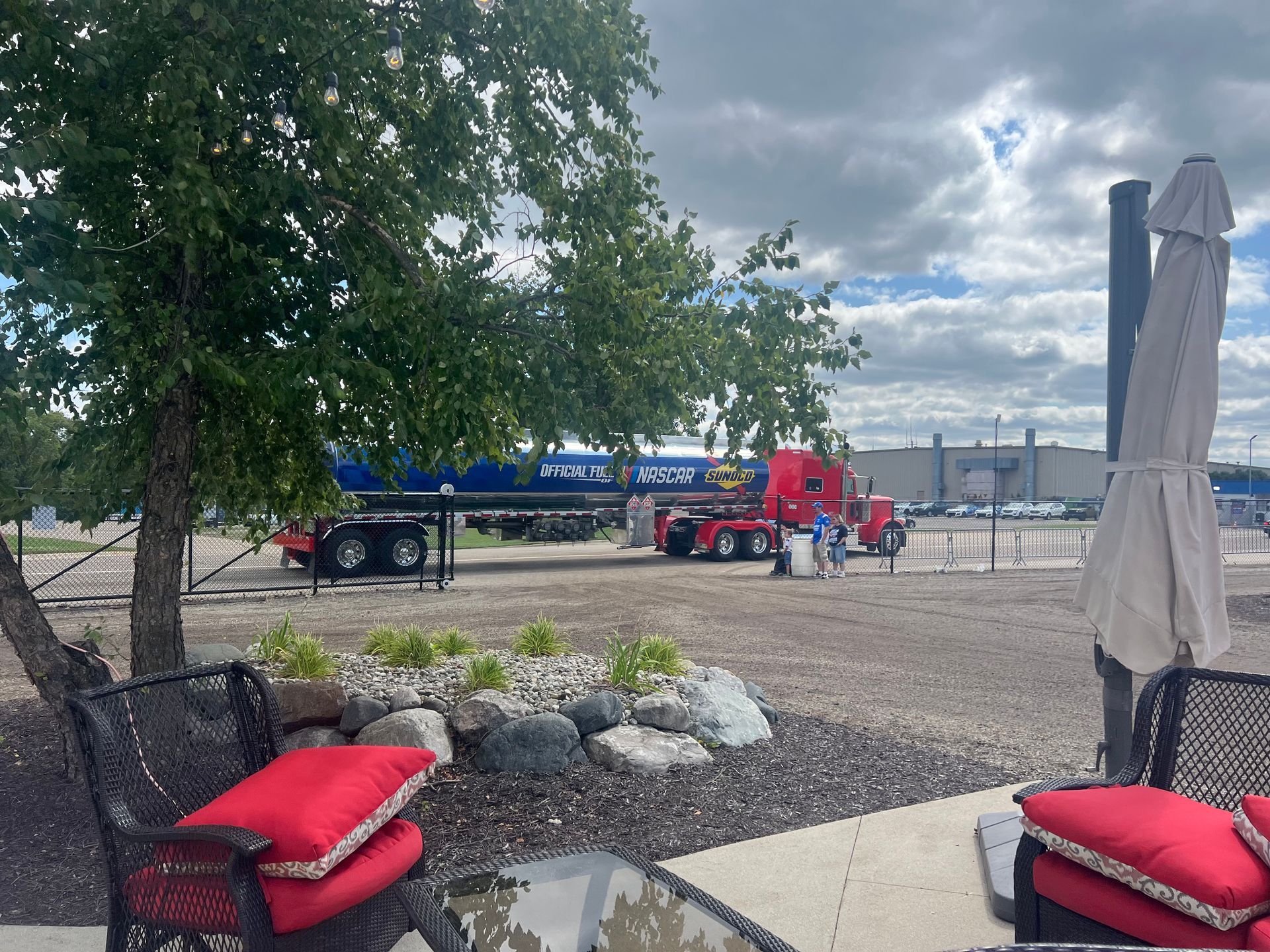 Red semi-truck with long trailer parked on gravel near a tree and outdoor seating area under a cloudy sky.
