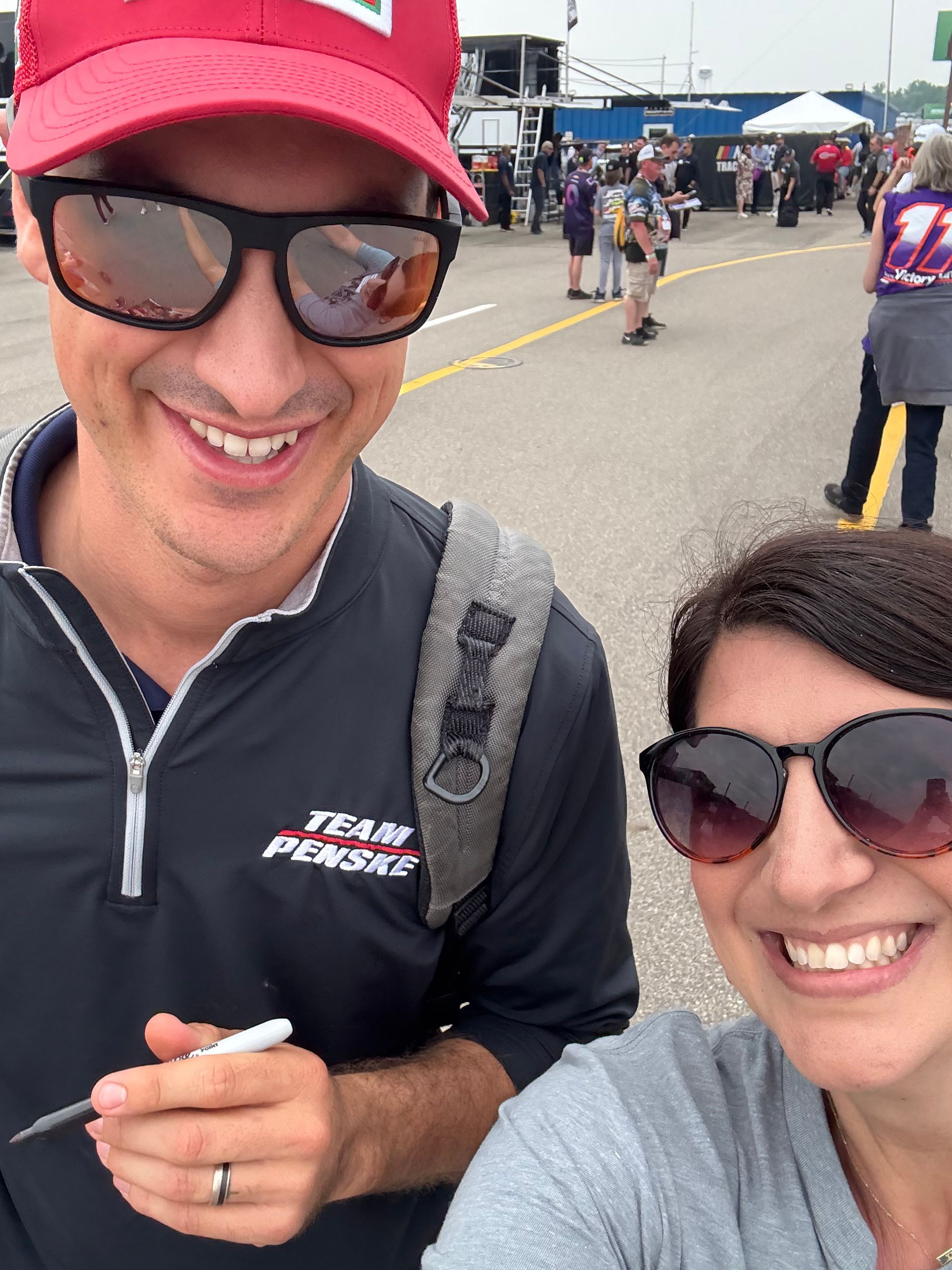Man in red cap and sunglasses smiles next to a woman wearing sunglasses. Both are outside, likely at a racetrack.