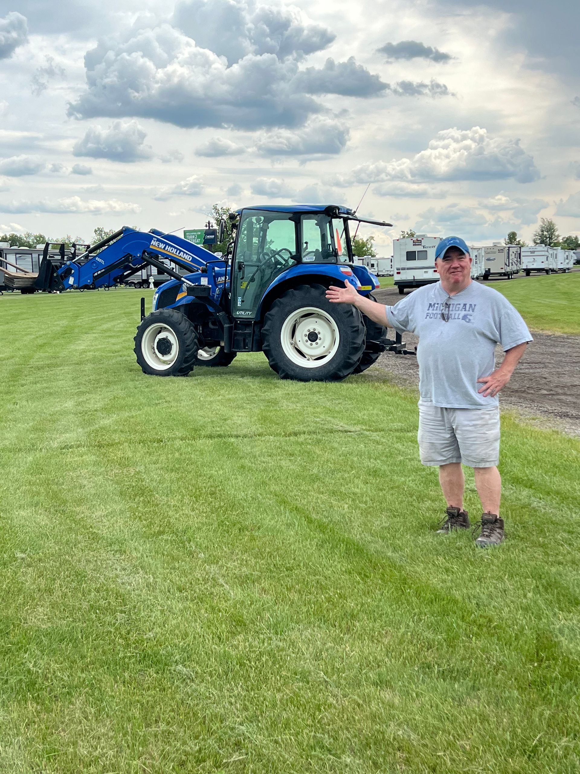 Man pointing at a blue tractor on a grassy field with RVs in the background, under a cloudy sky.