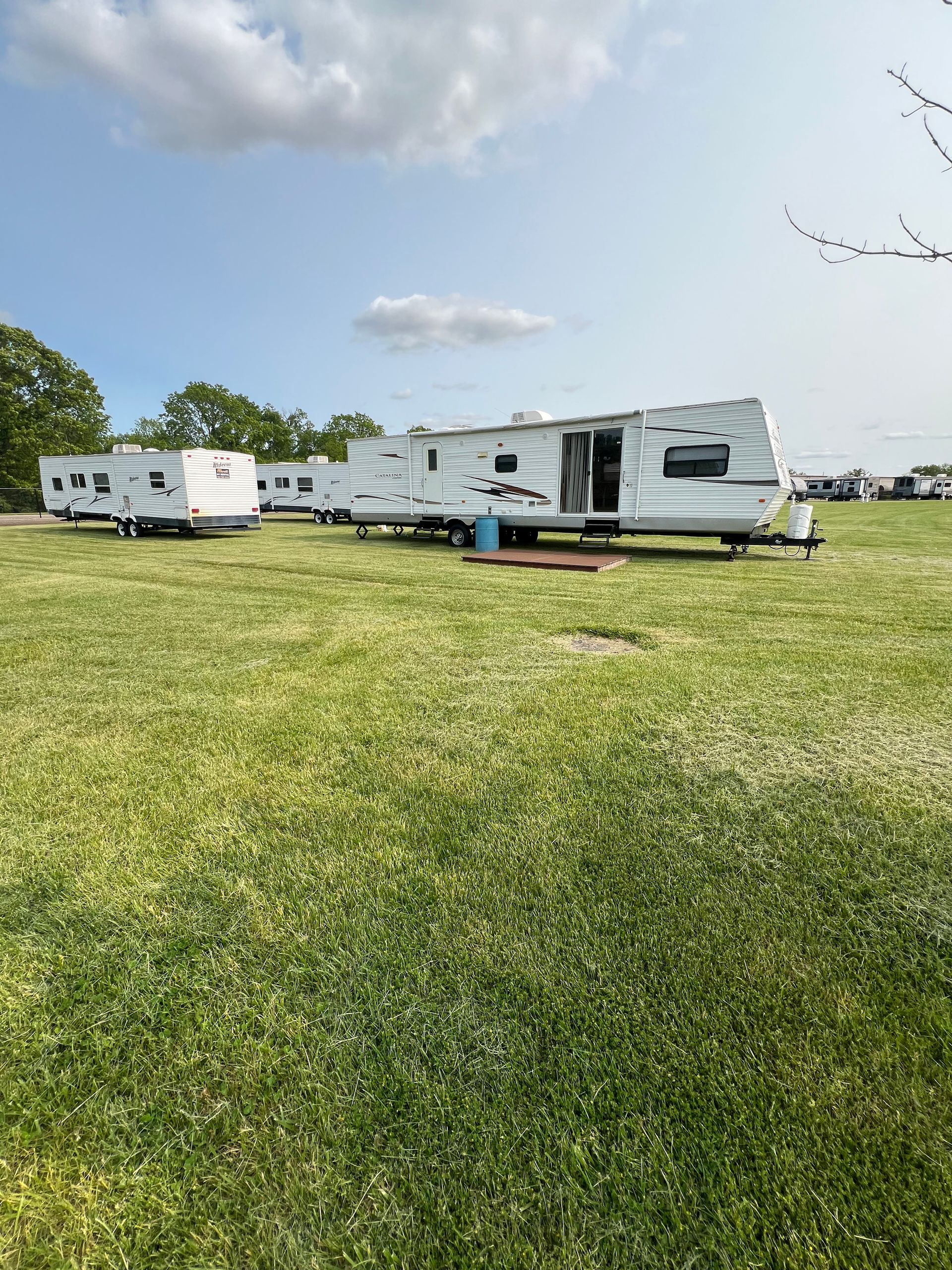 Campground scene: Several white RVs parked on a green grassy field under a cloudy blue sky.