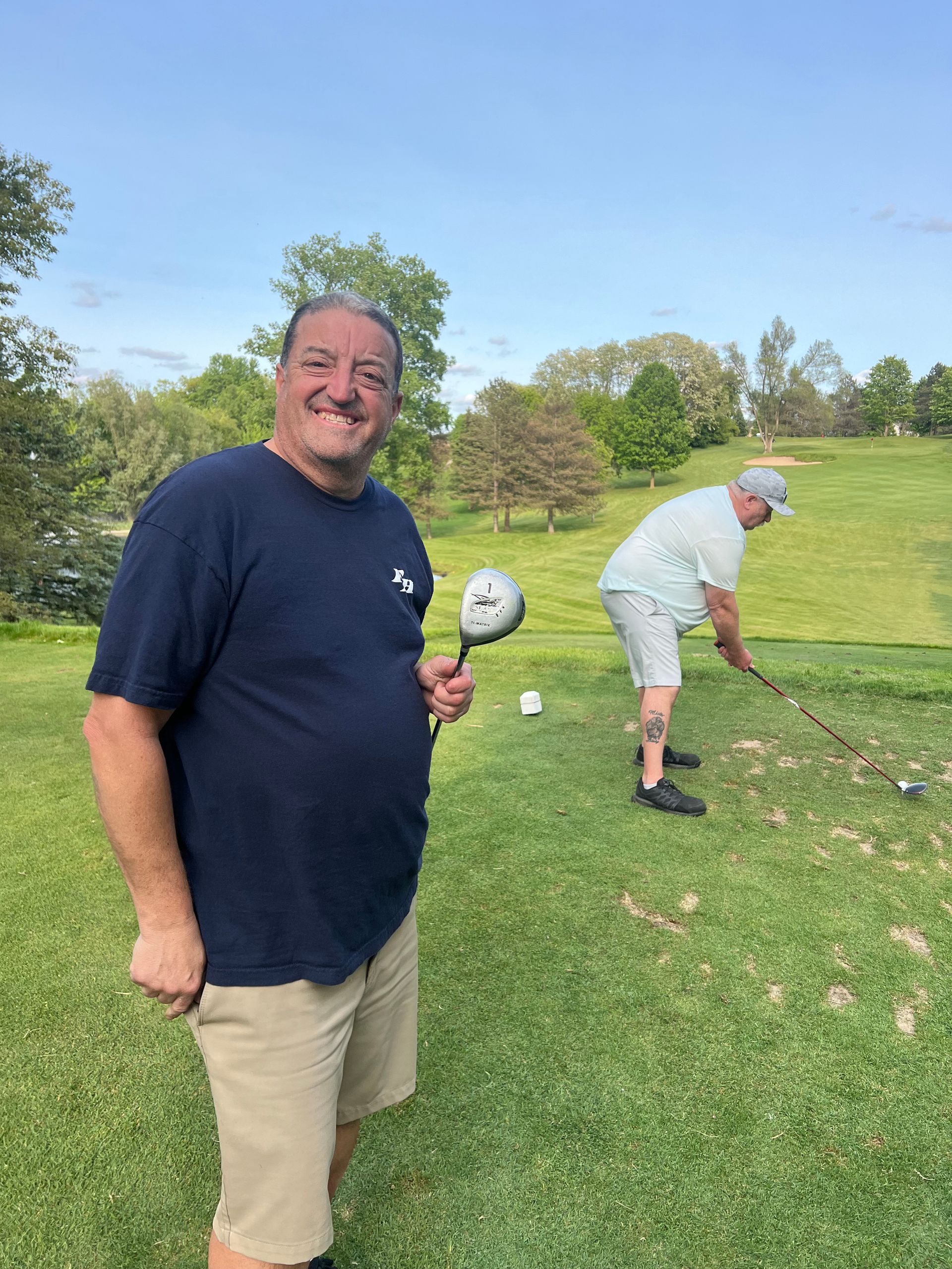 Man holding golf club, smiling on a green. Another golfer behind him is preparing to swing.