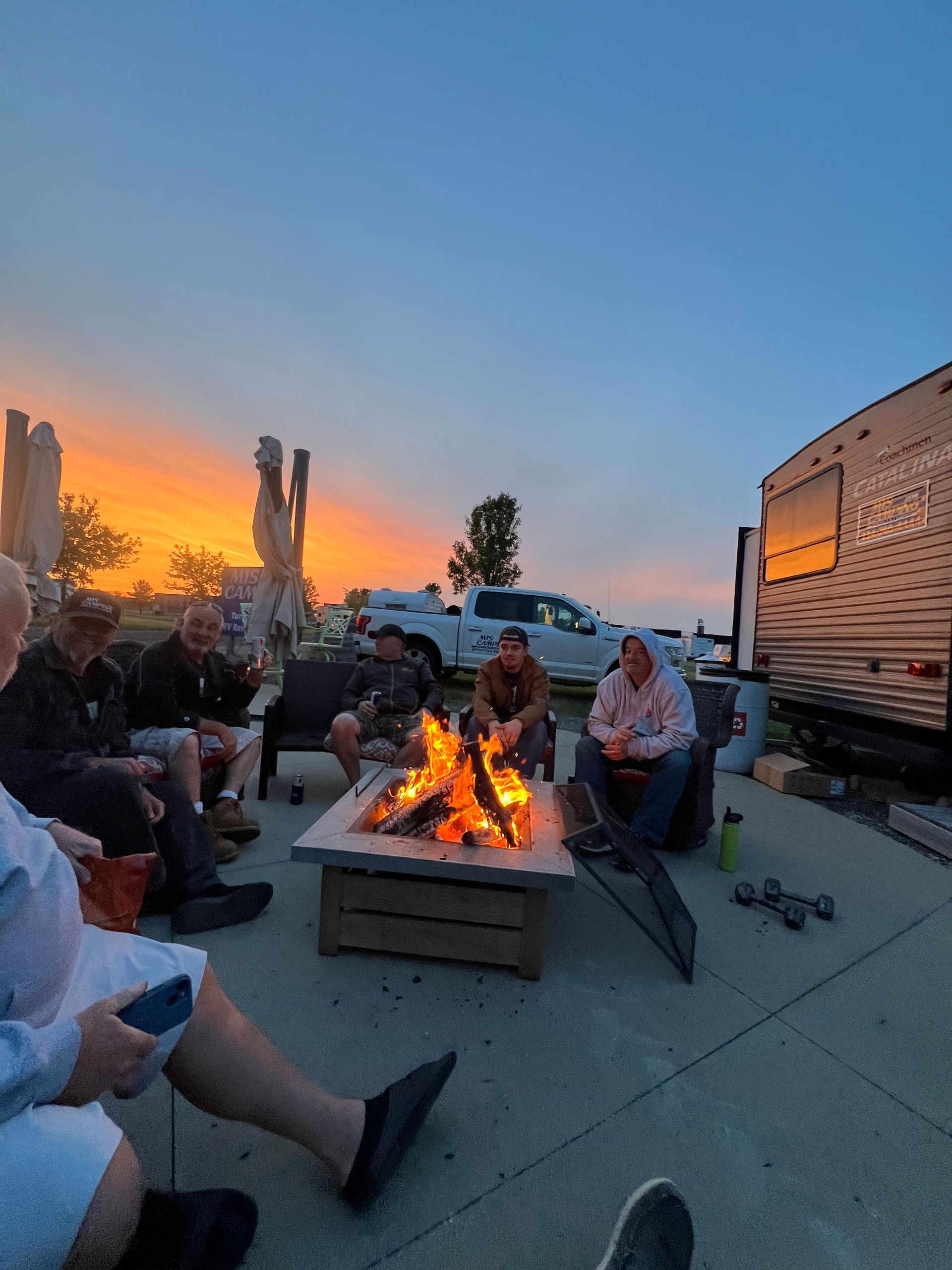 The Crew  around a campfire at sunset, with RVs and a truck in the background.