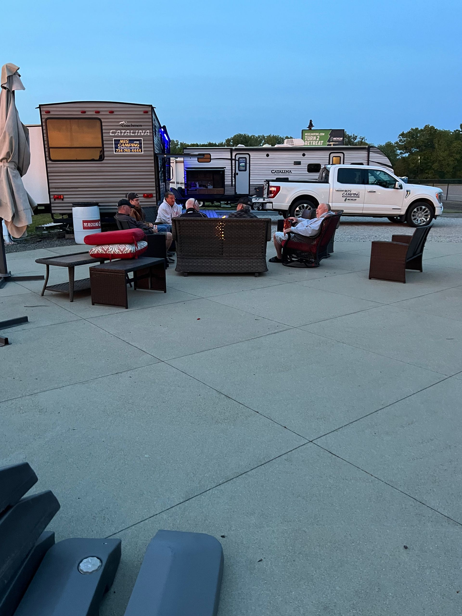 People sitting around a fire pit with trailers and a truck. Cloudy sky.