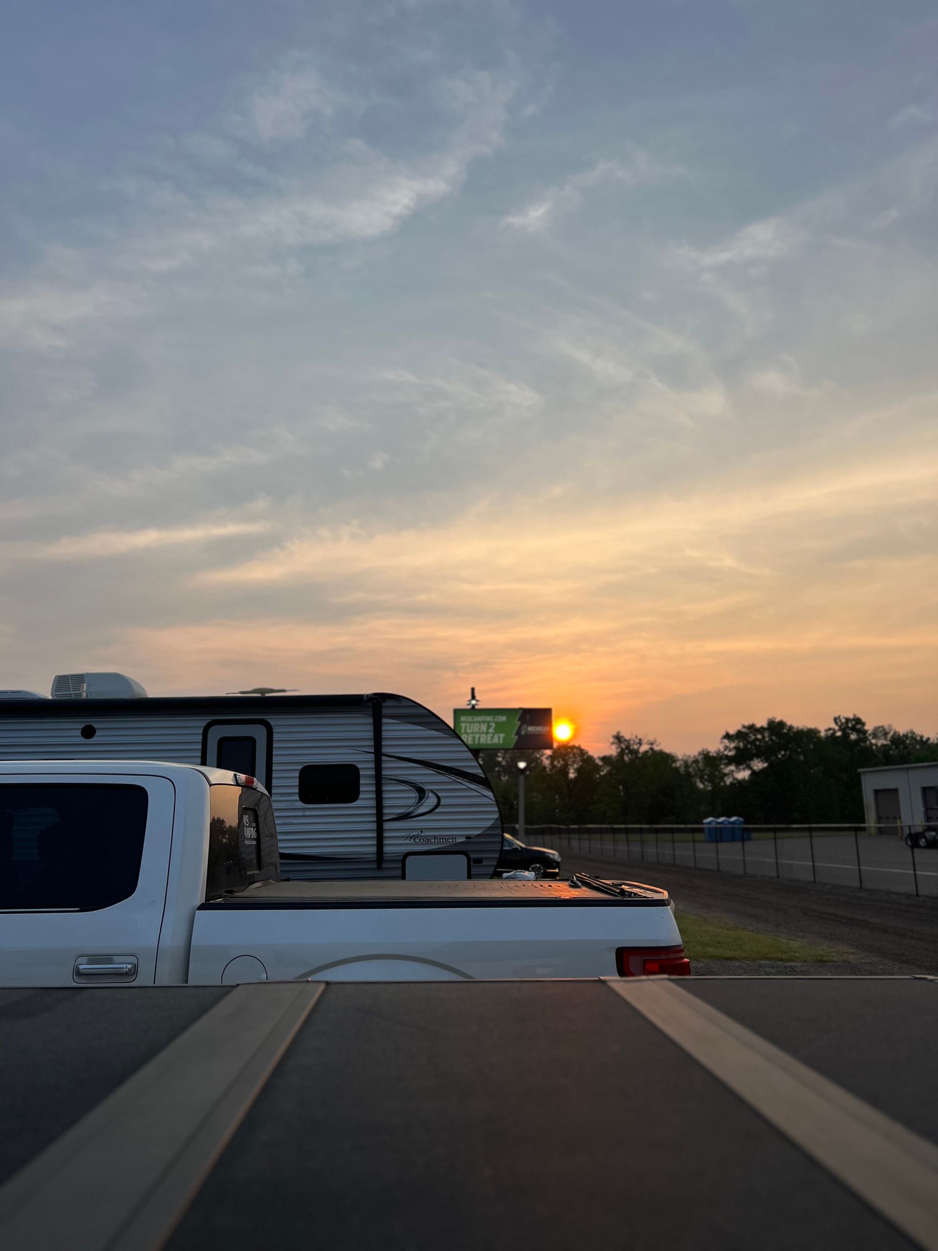 Sunset over parked RVs and a truck. Sky is pastel-colored; sun is peeking through trees.